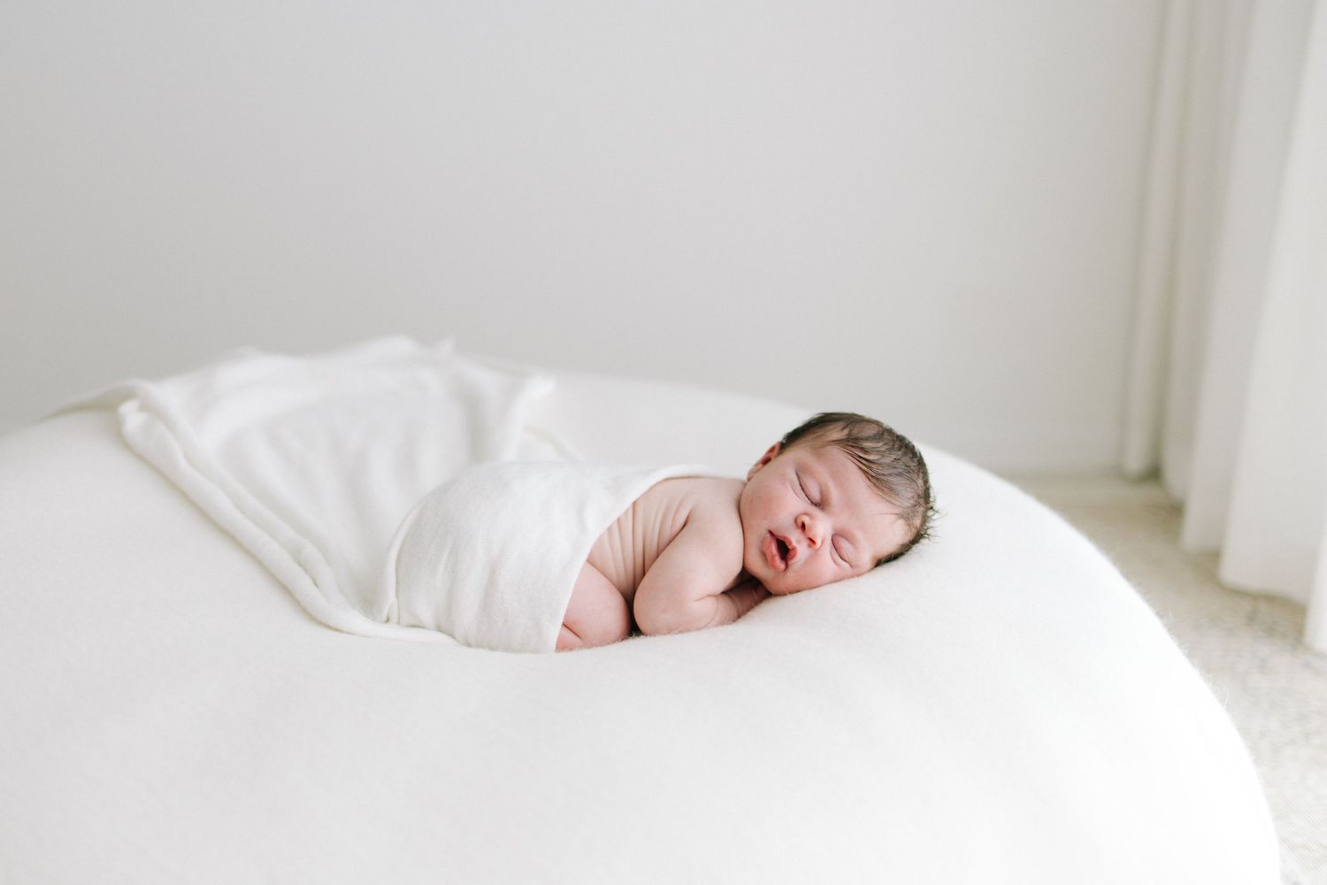 Swaddled newborn baby sleeping peacefully on a large white bean bag in bright room.
