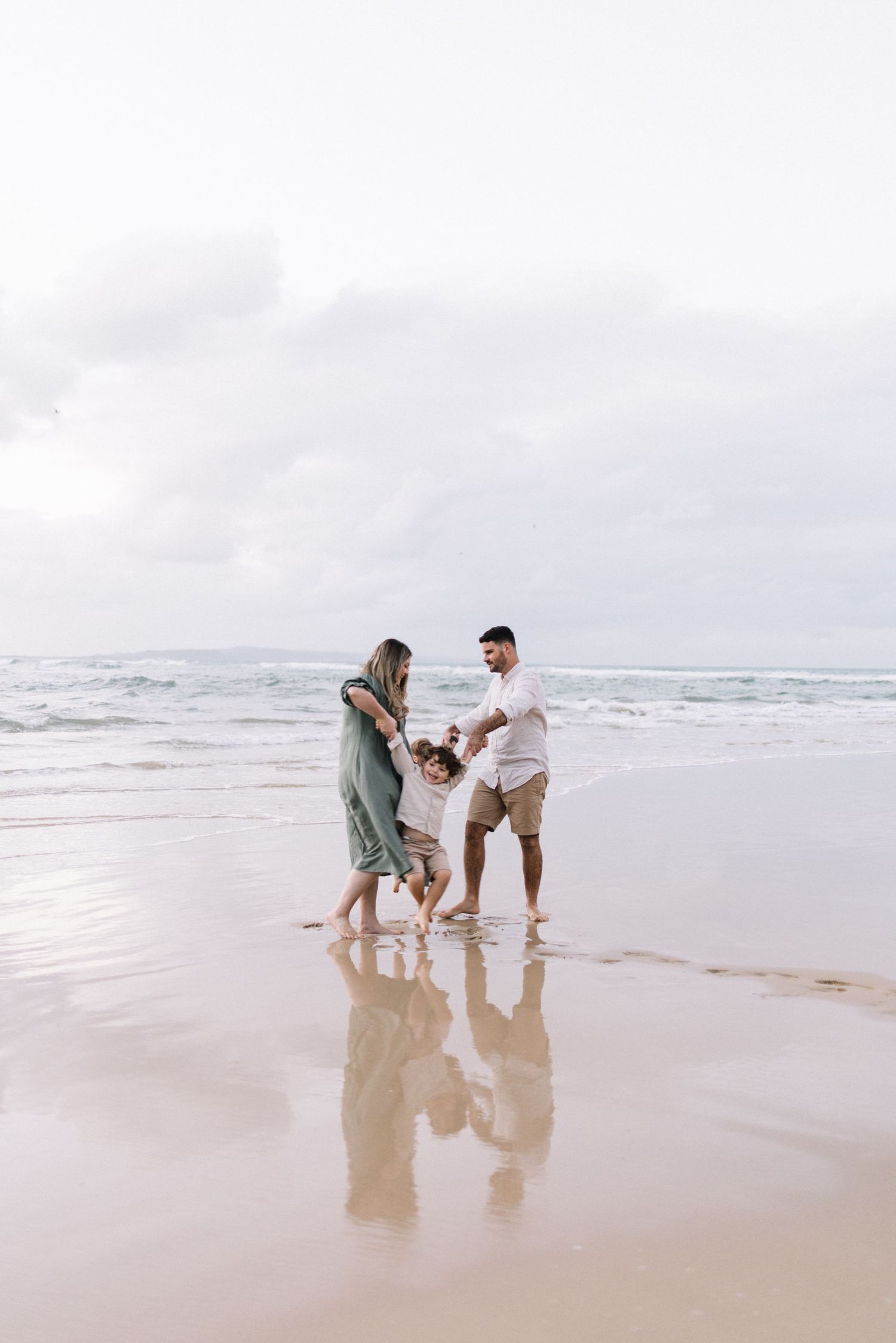 Happy family of three plays together on a reflective wet sandy beach with cloudy skies above.
