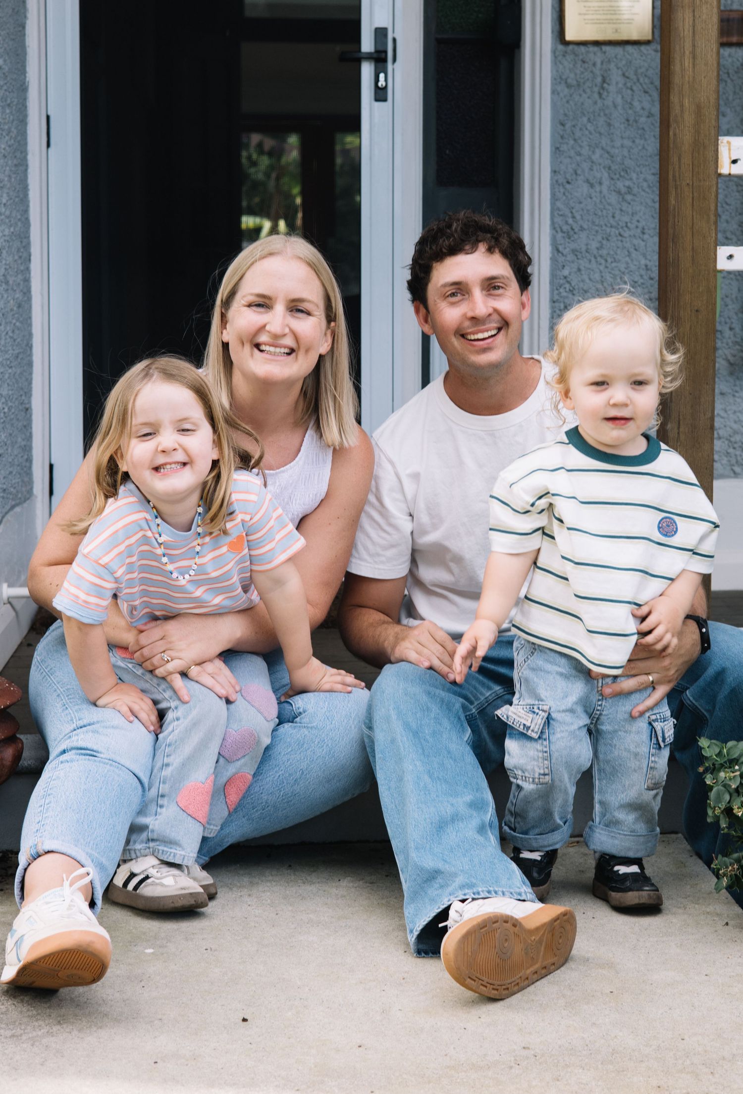 A happy young family of four smiling on front steps, two parents with a toddler boy and young girl in casual denim outfits.