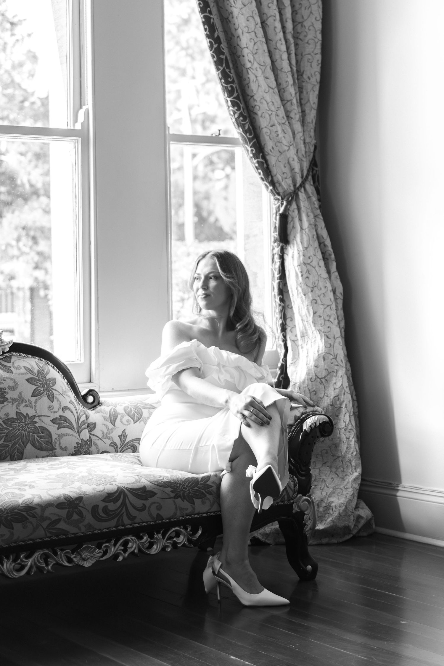 Black and white photo of elegant bride in white dress sitting on ornate chaise lounge by large window with drapes.