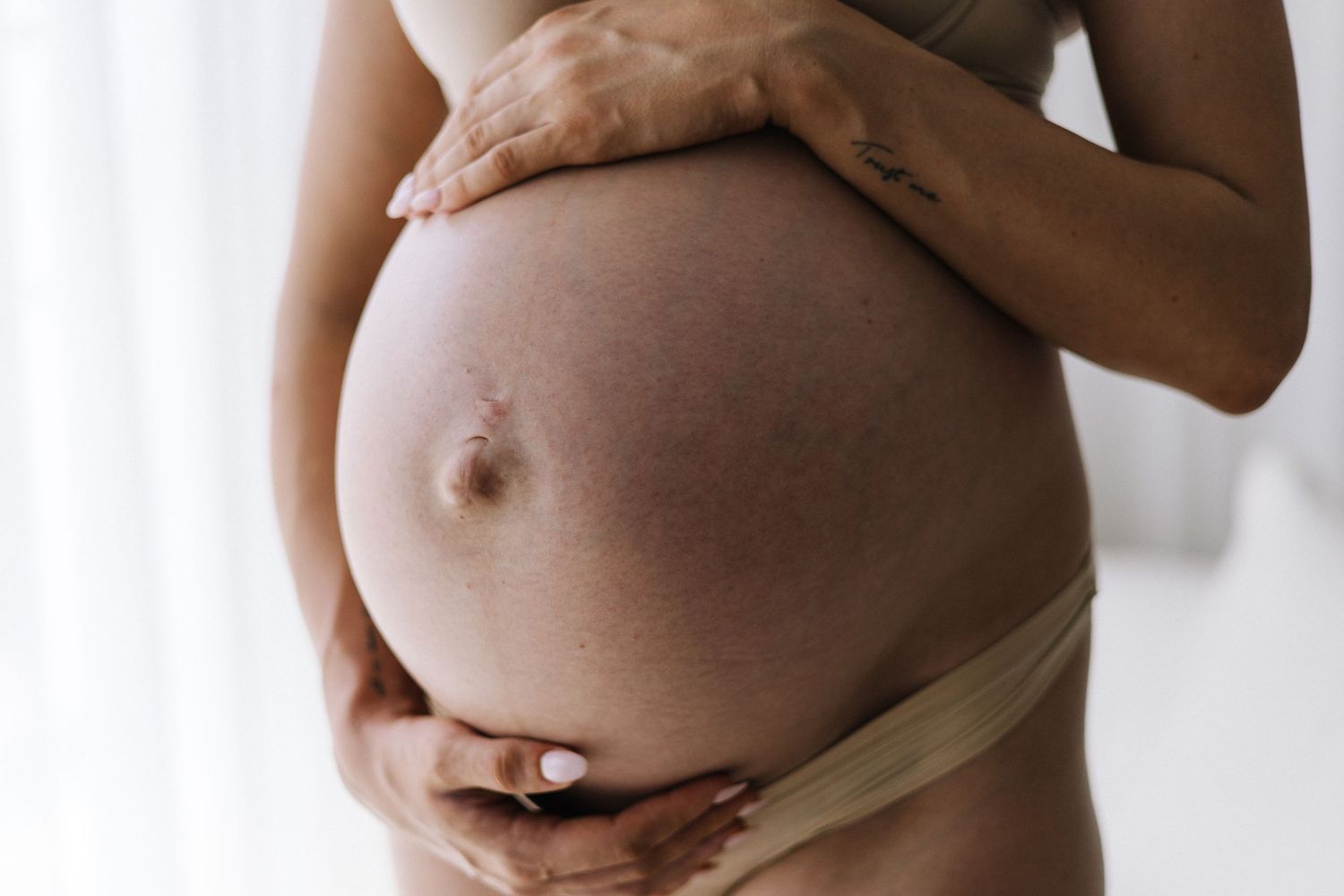 Close-up of a pregnant woman cradling her bare baby bump with both hands against a soft white background.