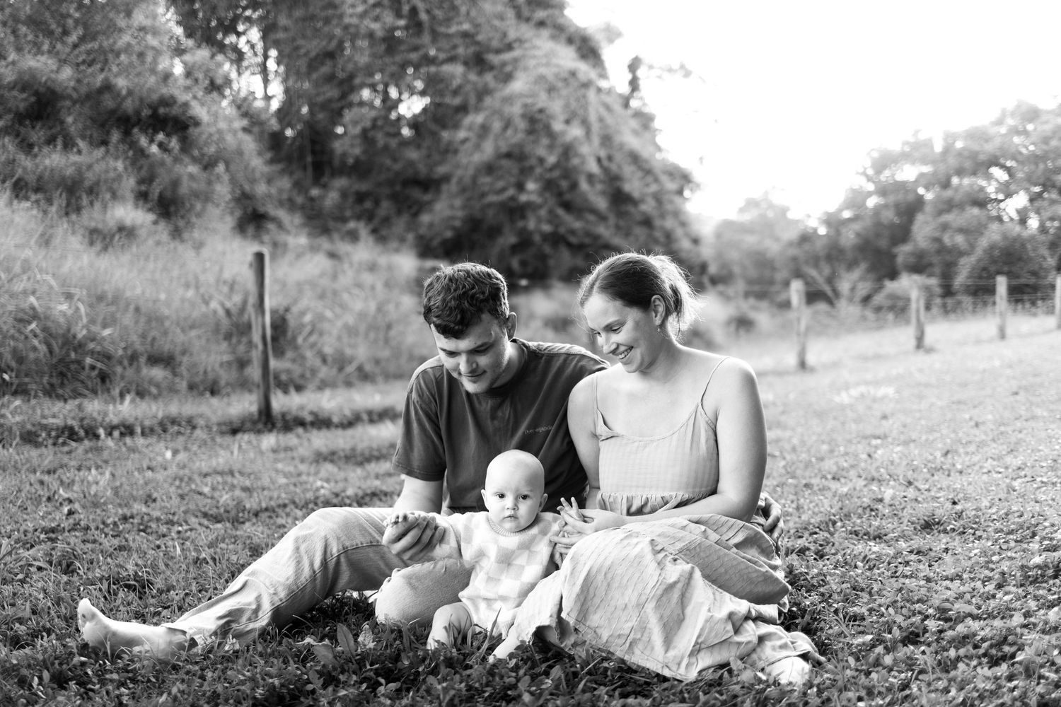 Black and white photo of a young family sitting on grass outdoors, parents smiling at their baby.