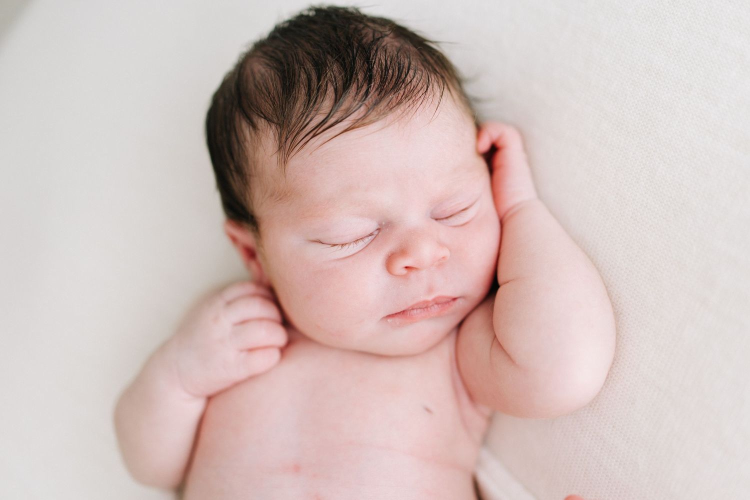 Peaceful sleeping newborn baby with dark hair, hands tucked under cheeks, lying on soft white background.