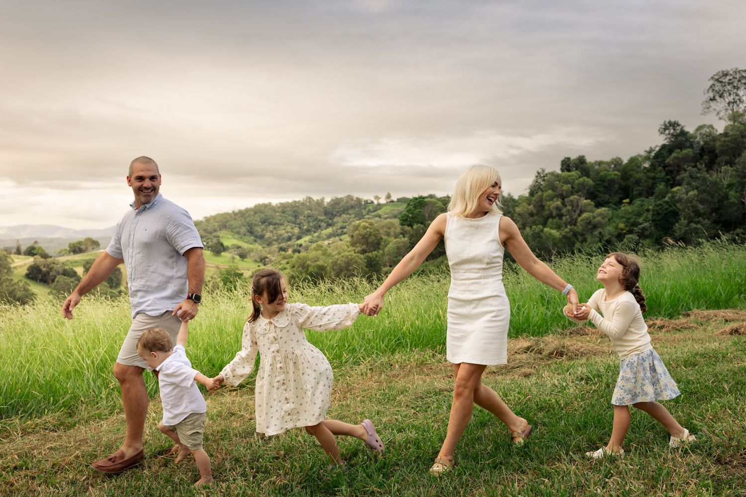 A happy family of four holds hands and dances together in a scenic green meadow at sunset.