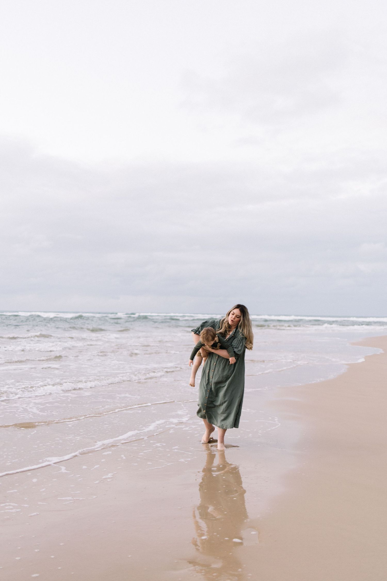 A woman in a green maxi dress laughs while carrying a young child along a wet sandy beach shoreline.