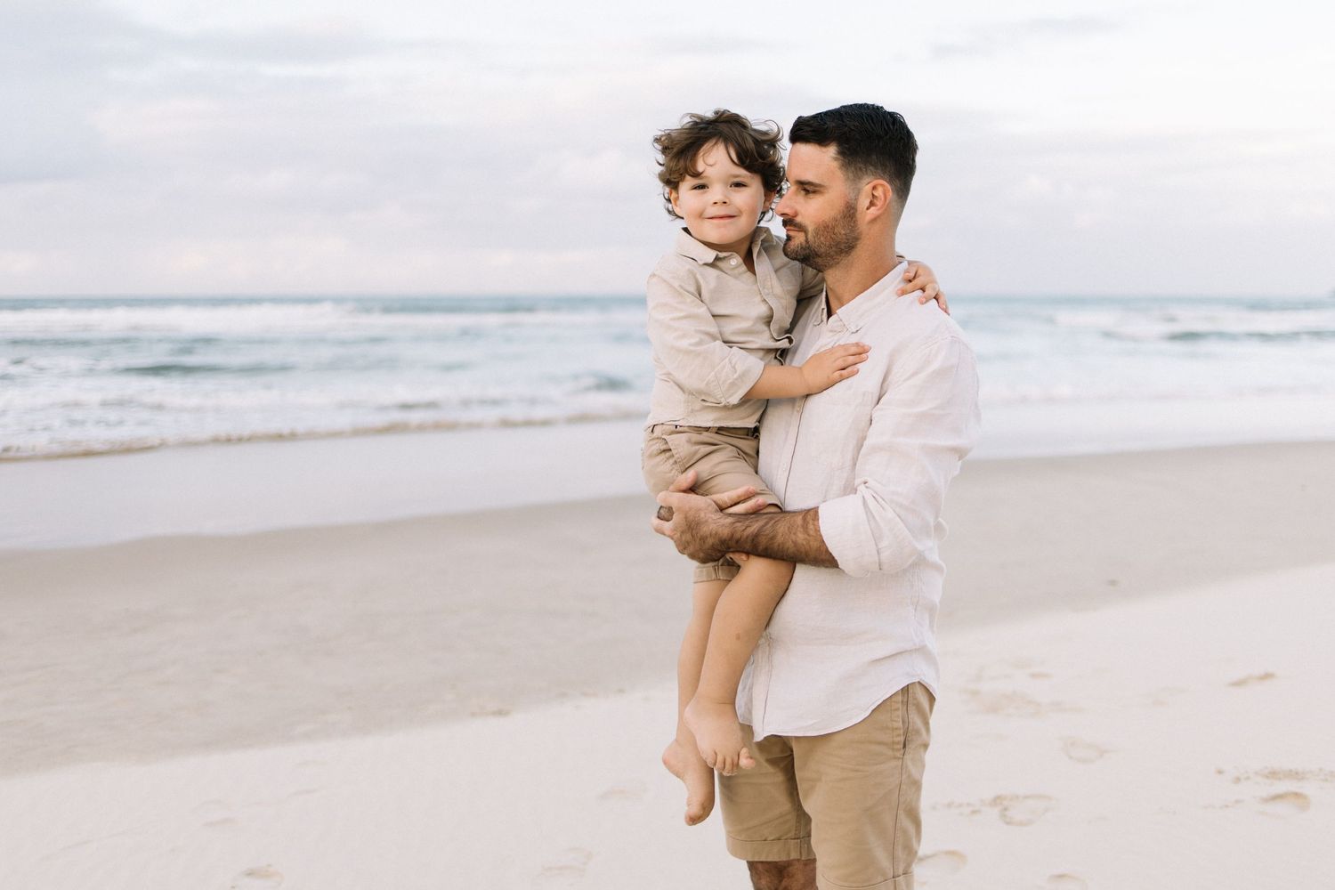 A father holds his young son on a sandy beach, sharing a tender moment with ocean waves in the background.