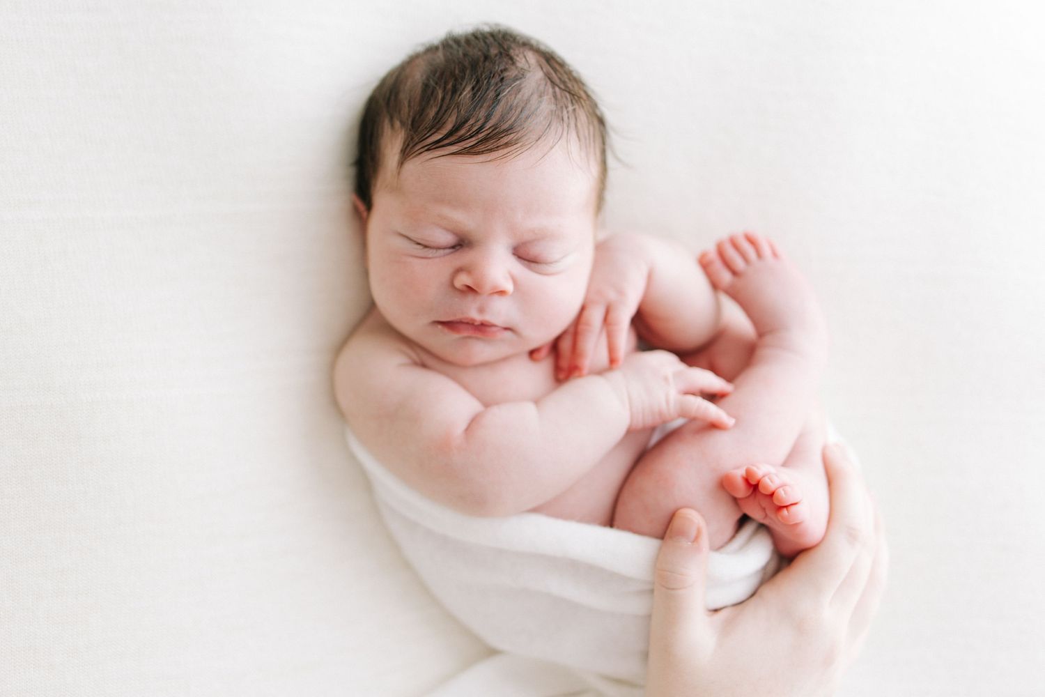 Peaceful newborn baby sleeping wrapped in white cloth, cradled in parent's hands on soft white background.