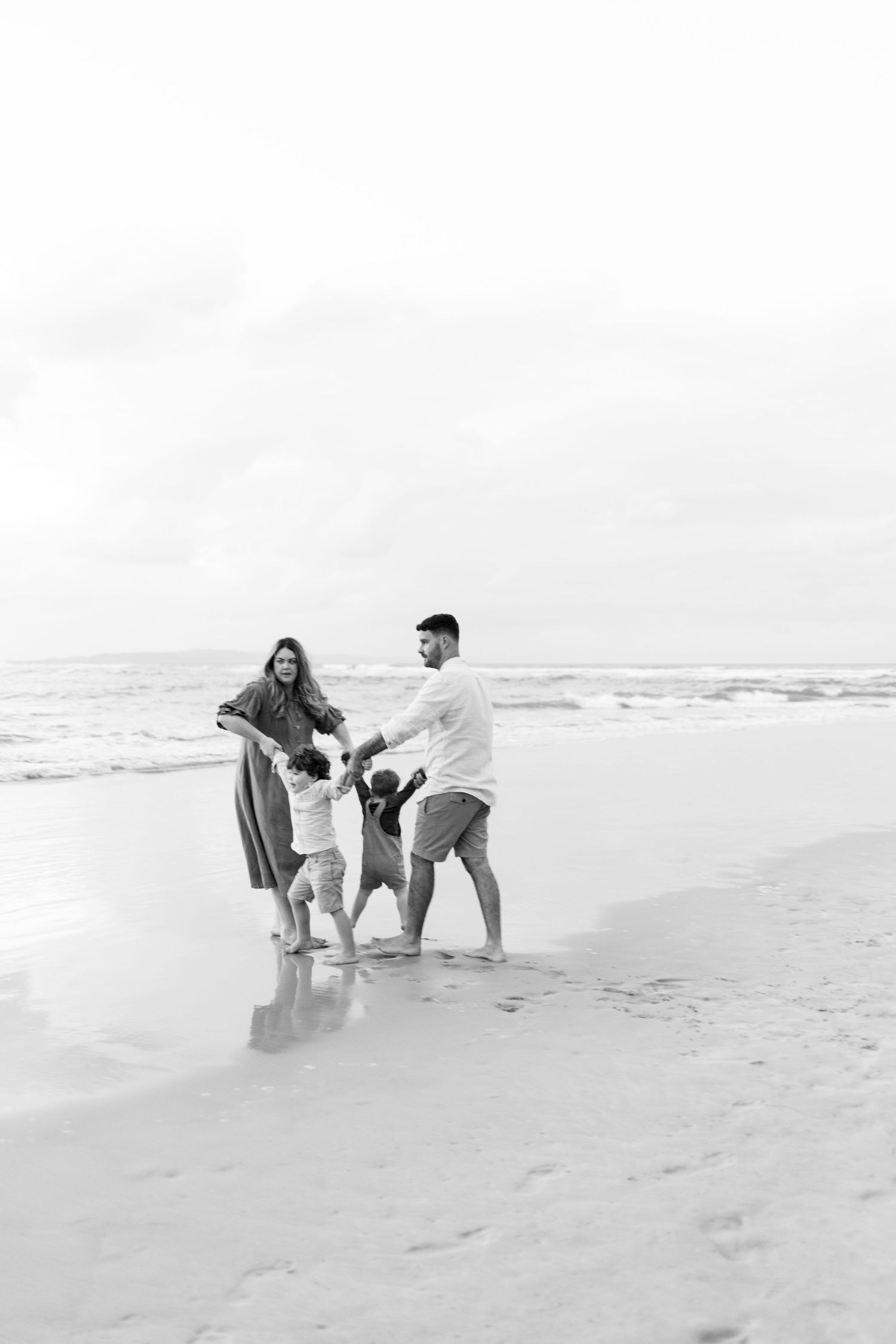 Black and white photo of a family of four holding hands and playing together on a beach.