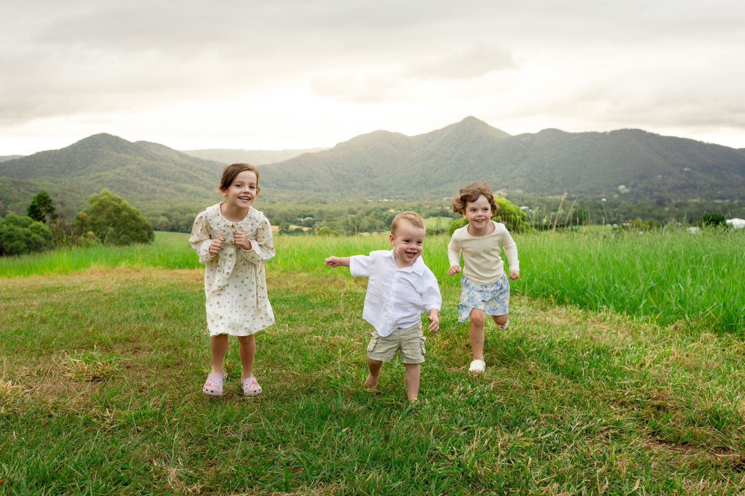 Three young children laugh and run through a lush green field with mountain views in the background.