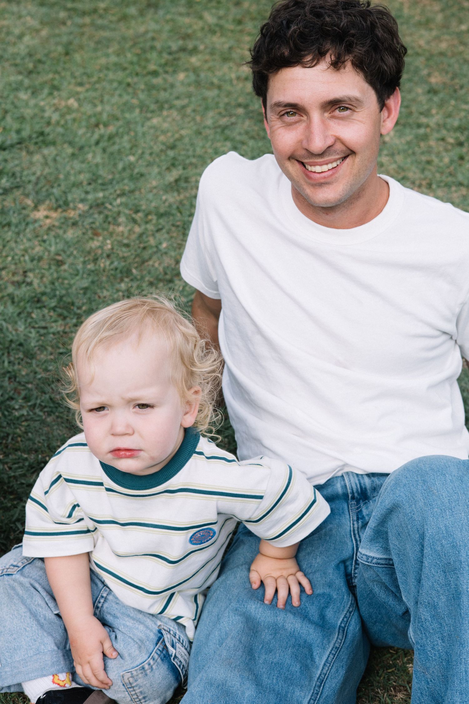 Smiling man in white t-shirt sitting on grass with a blonde toddler in striped shirt and jeans.
