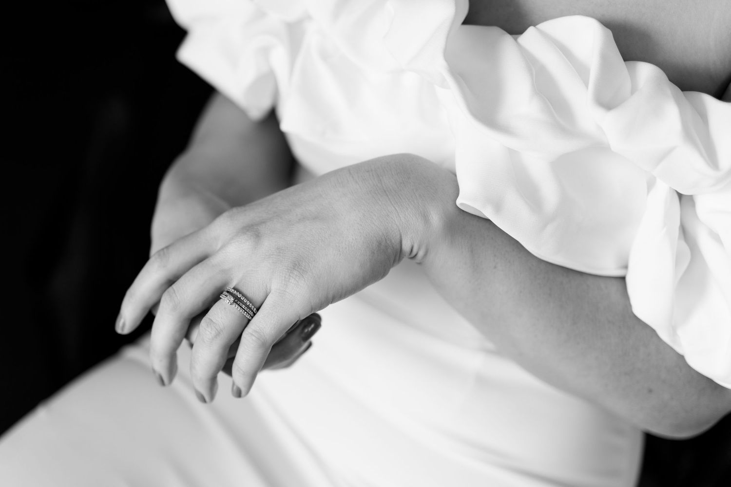 Black and white close-up of a bride's hand wearing a diamond ring, resting on a white wedding dress.