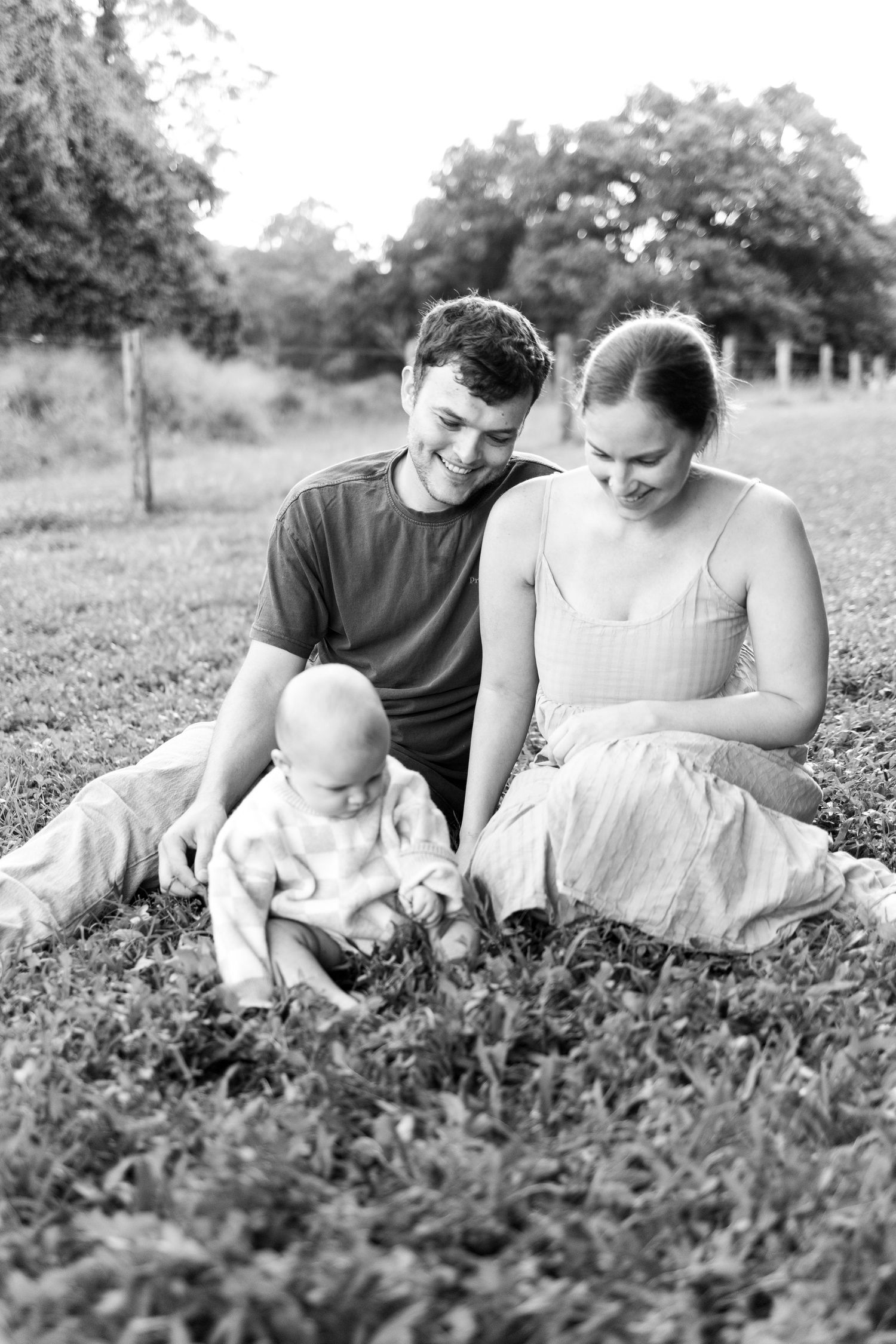 Black and white photo of a young family sitting on grass, parents smiling at their baby exploring the ground.