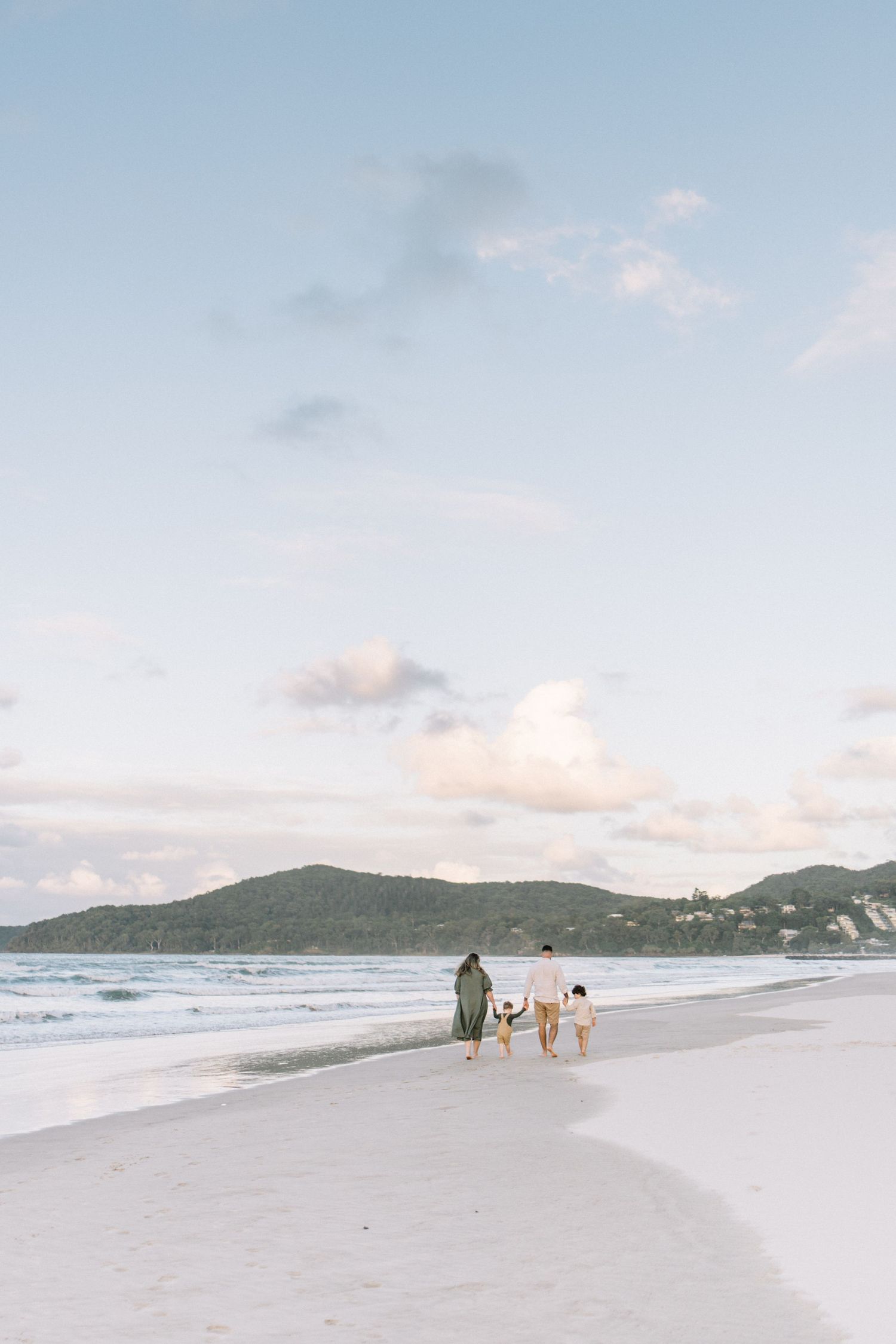 Family walking along a white sandy beach at sunset with green hills and ocean waves in the background.
