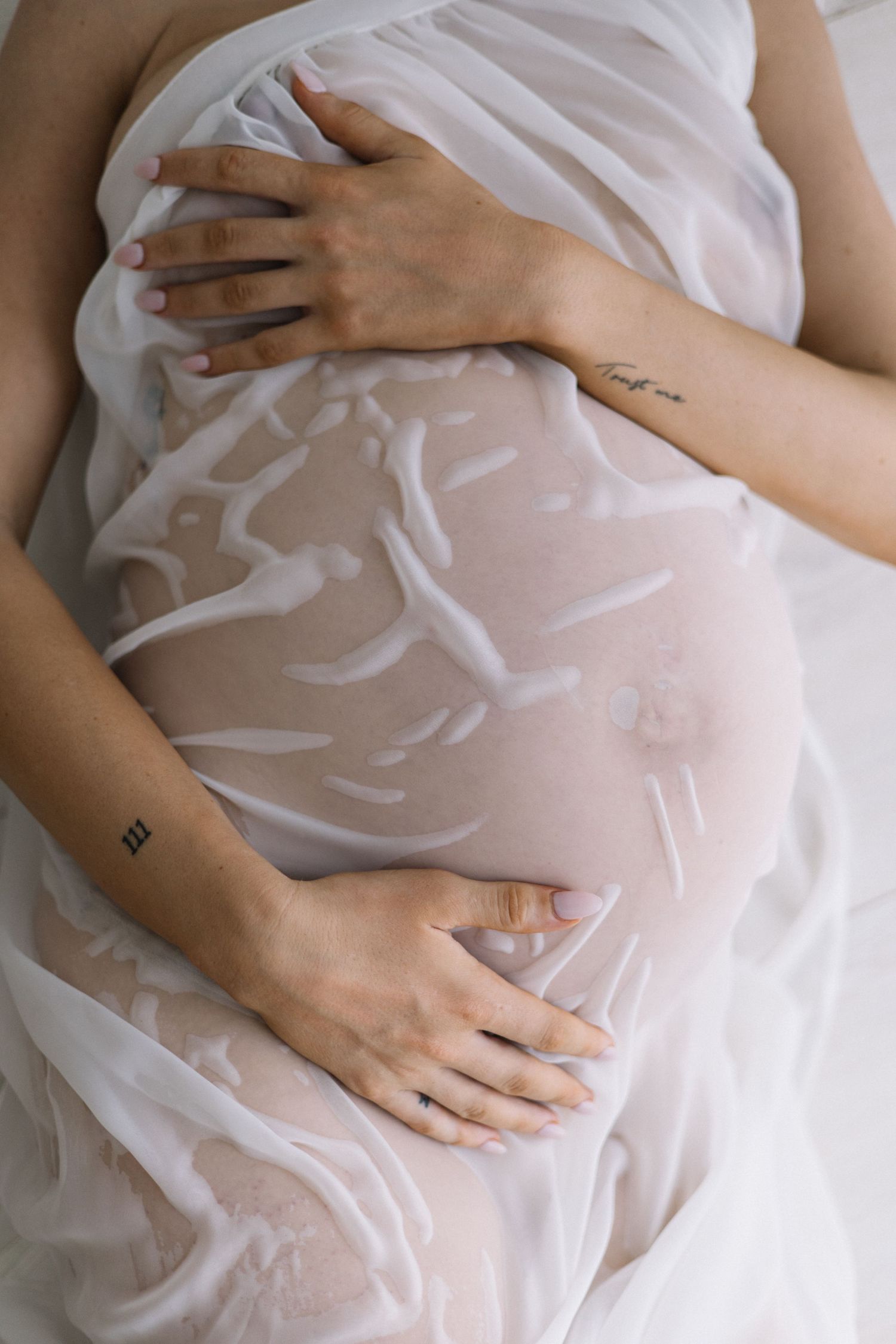Close-up of pregnant belly wrapped in sheer white fabric with tattooed hands gently cradling bump.