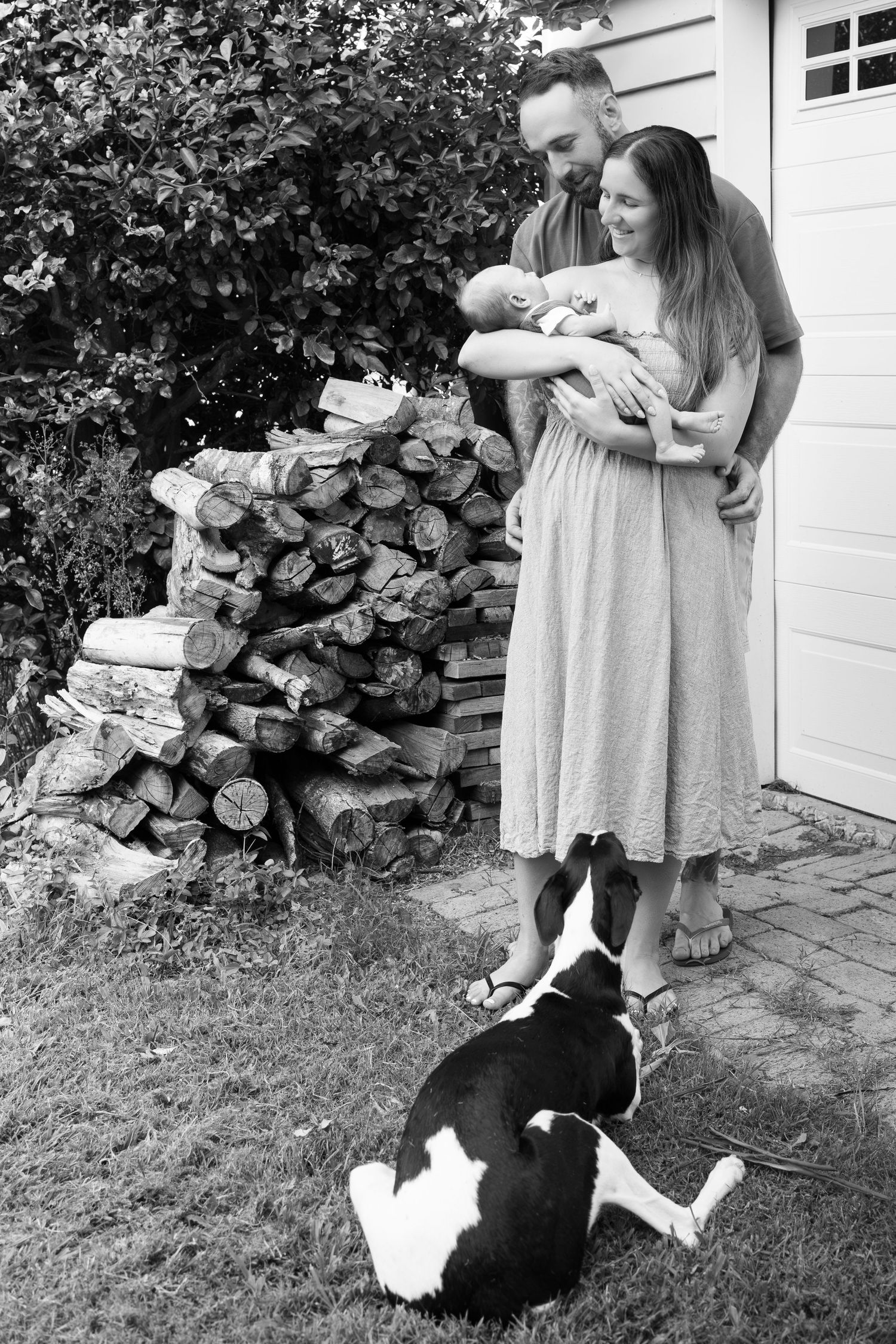 Happy couple admires their newborn baby outdoors as a black and white dog watches near a woodpile.