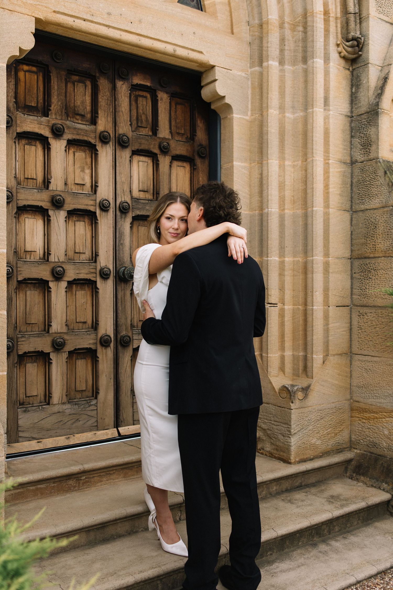 A couple embraces in wedding attire before ornate wooden doors of a stone church, bride in white dress.