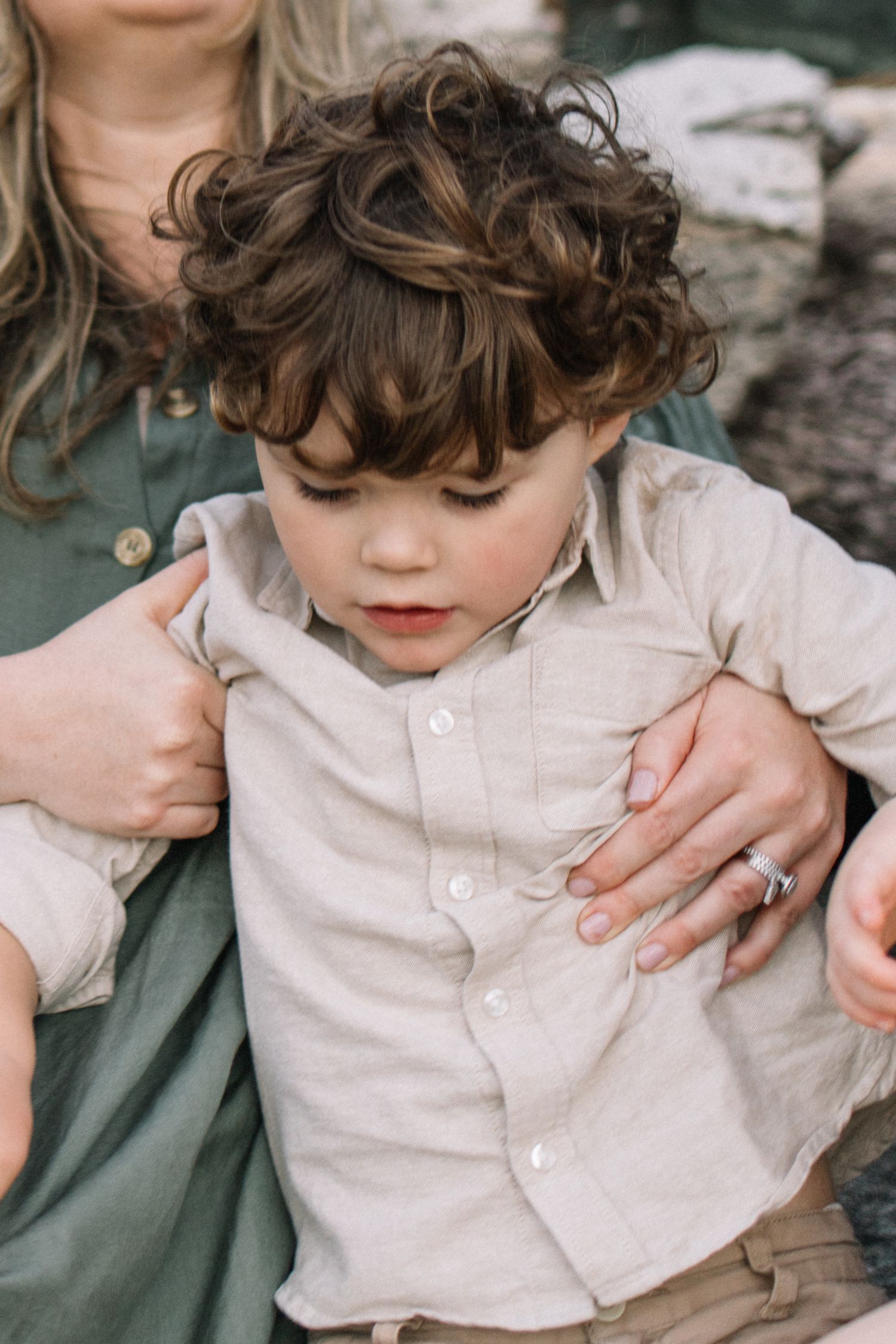 Curly-haired toddler boy in a beige linen shirt sitting with his mother outdoors.