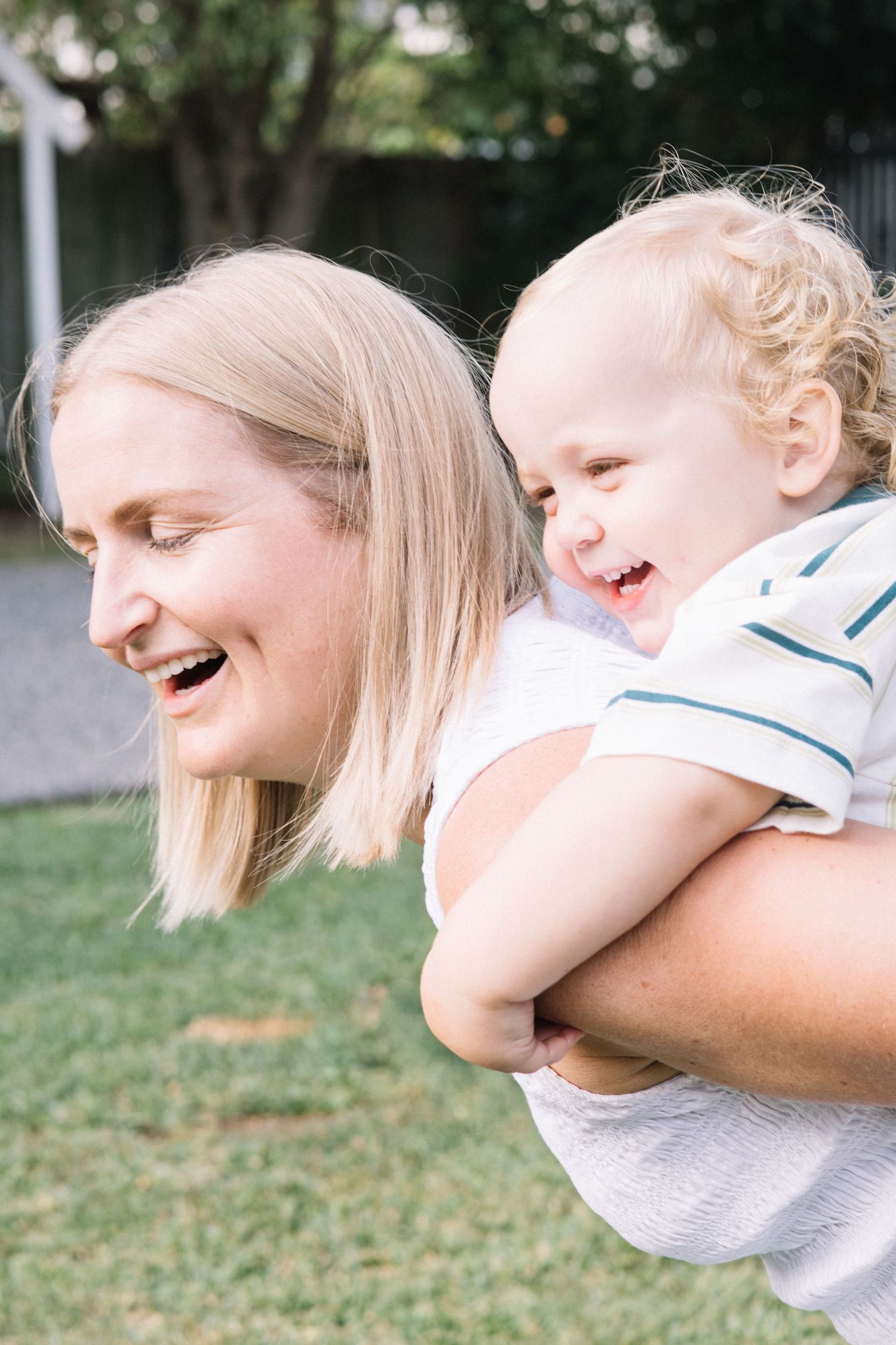 A laughing blonde woman gives a giggling toddler boy a piggyback ride outdoors on a sunny day.