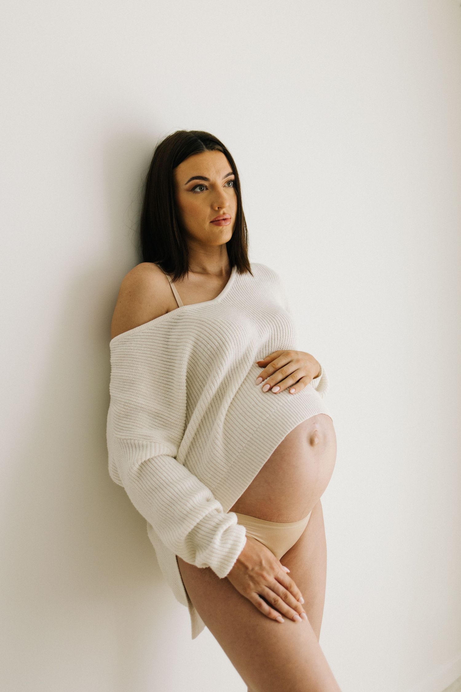 Pregnant woman in white off-shoulder sweater and nude underwear cradling baby bump against white wall.