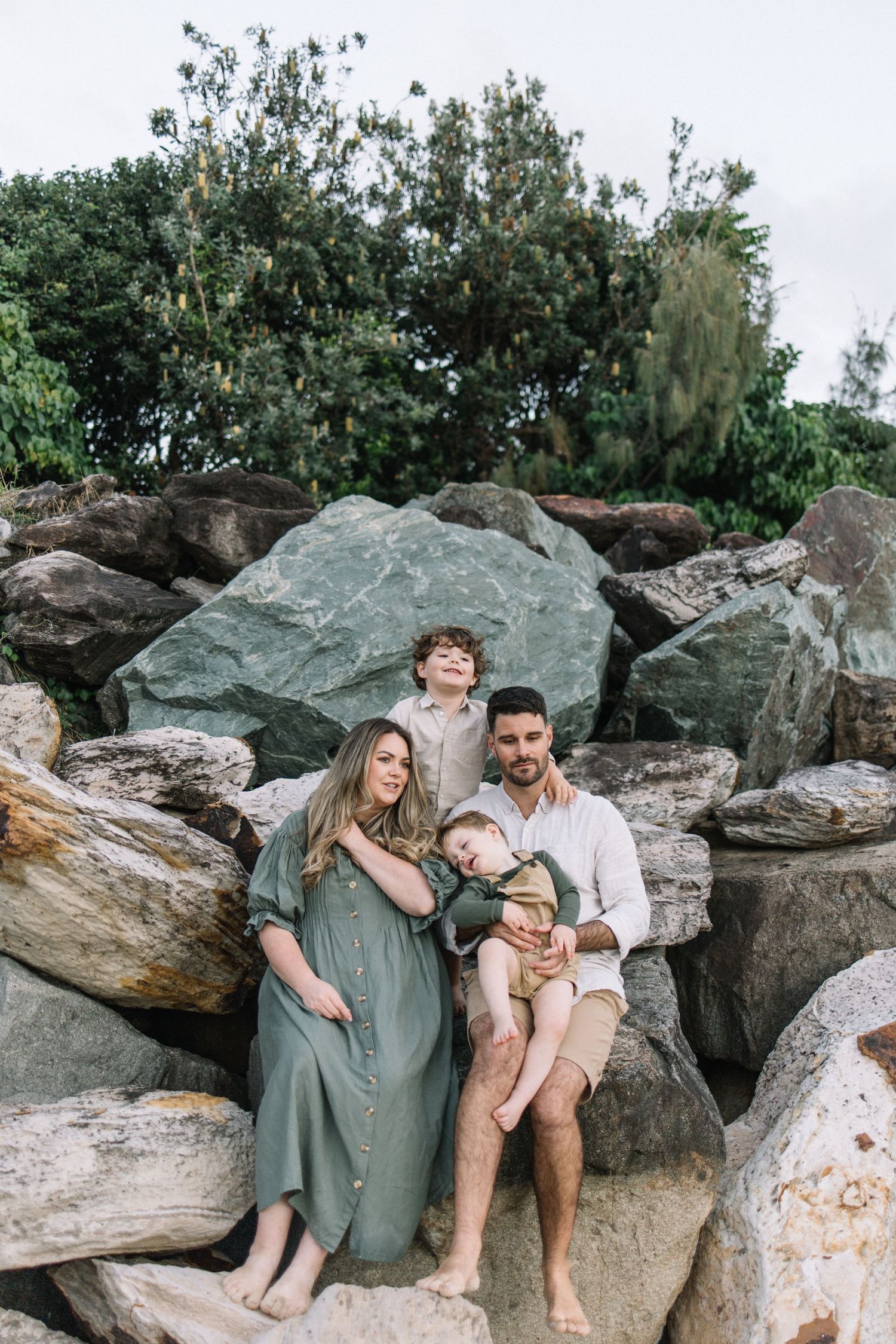 Family of four sitting on coastal rocks, mother in green dress with two young boys and father in white shirt.