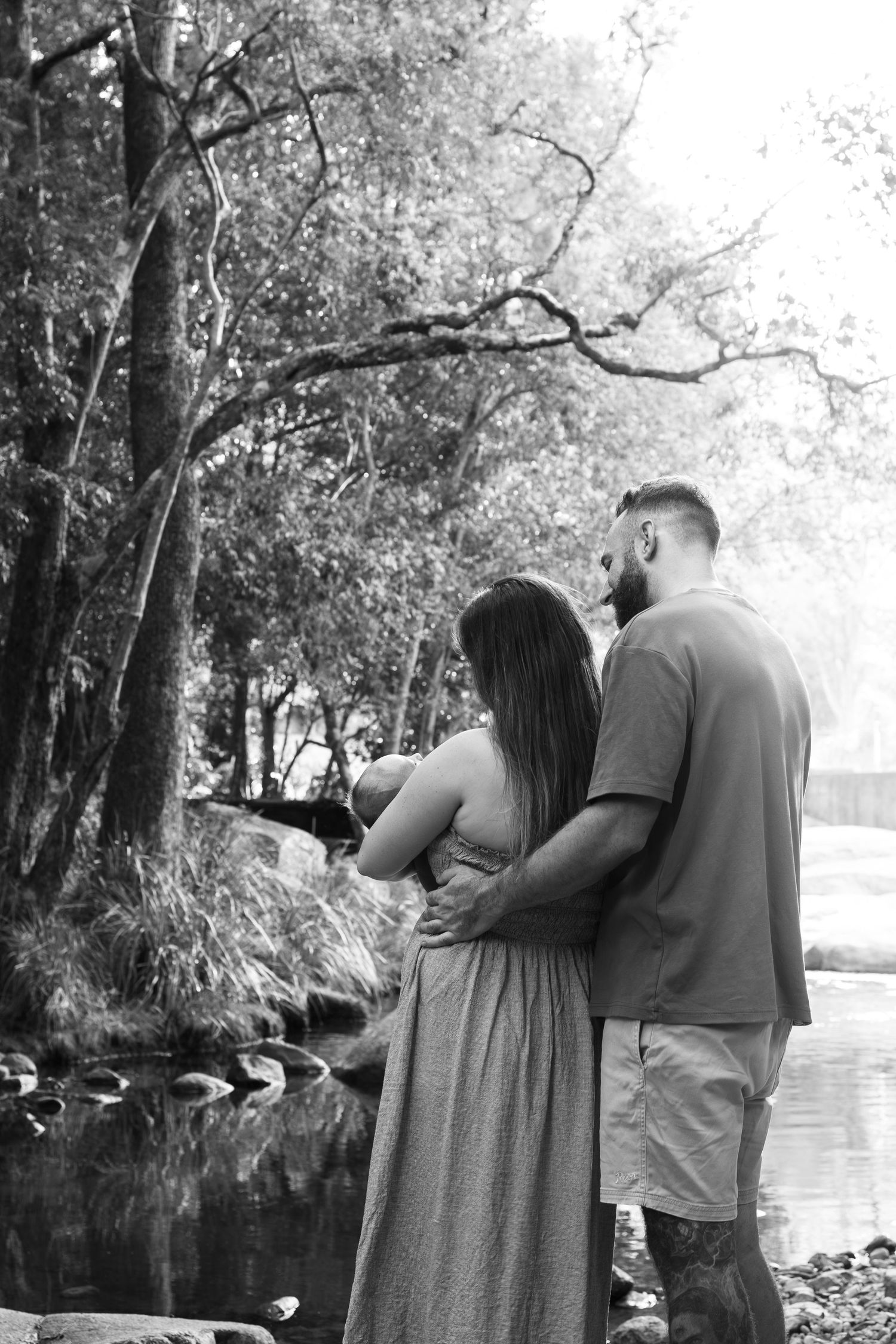 Black and white photo of a couple embracing by a serene forest creek, gazing at the water surrounded by trees.