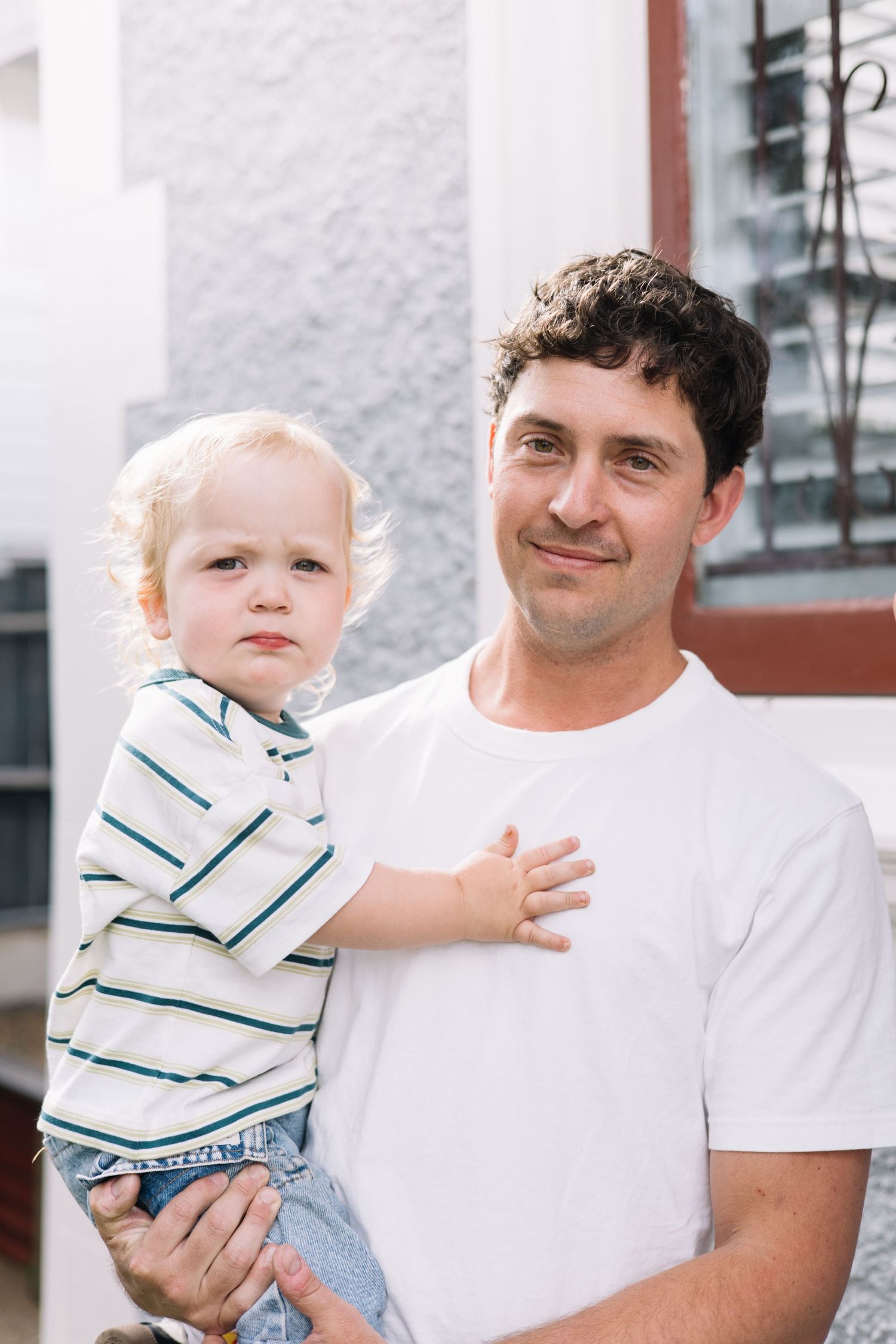 A smiling man in a white t-shirt holds a young blonde toddler in a striped shirt outdoors near a building.