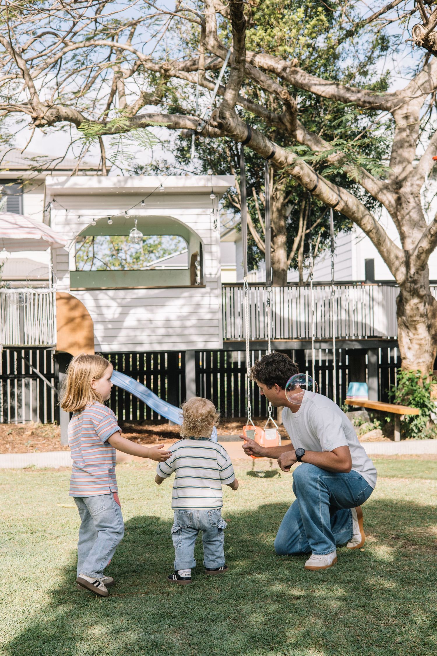 Family playing outdoors in backyard with treehouse, parent crouching to interact with two young children on green lawn.