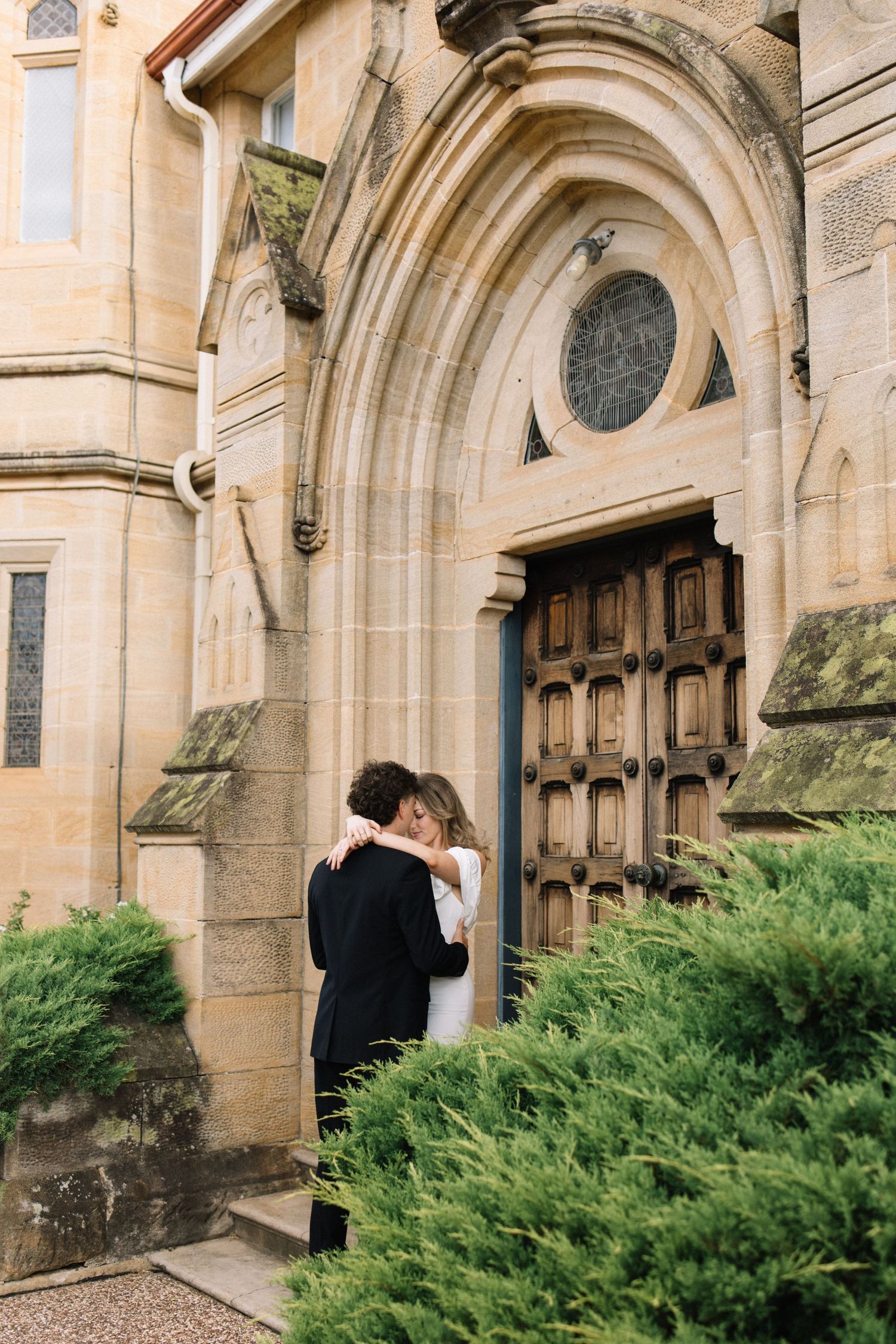 Couple embracing in wedding attire before ornate stone church entrance with arched doorway and lush green shrubs.