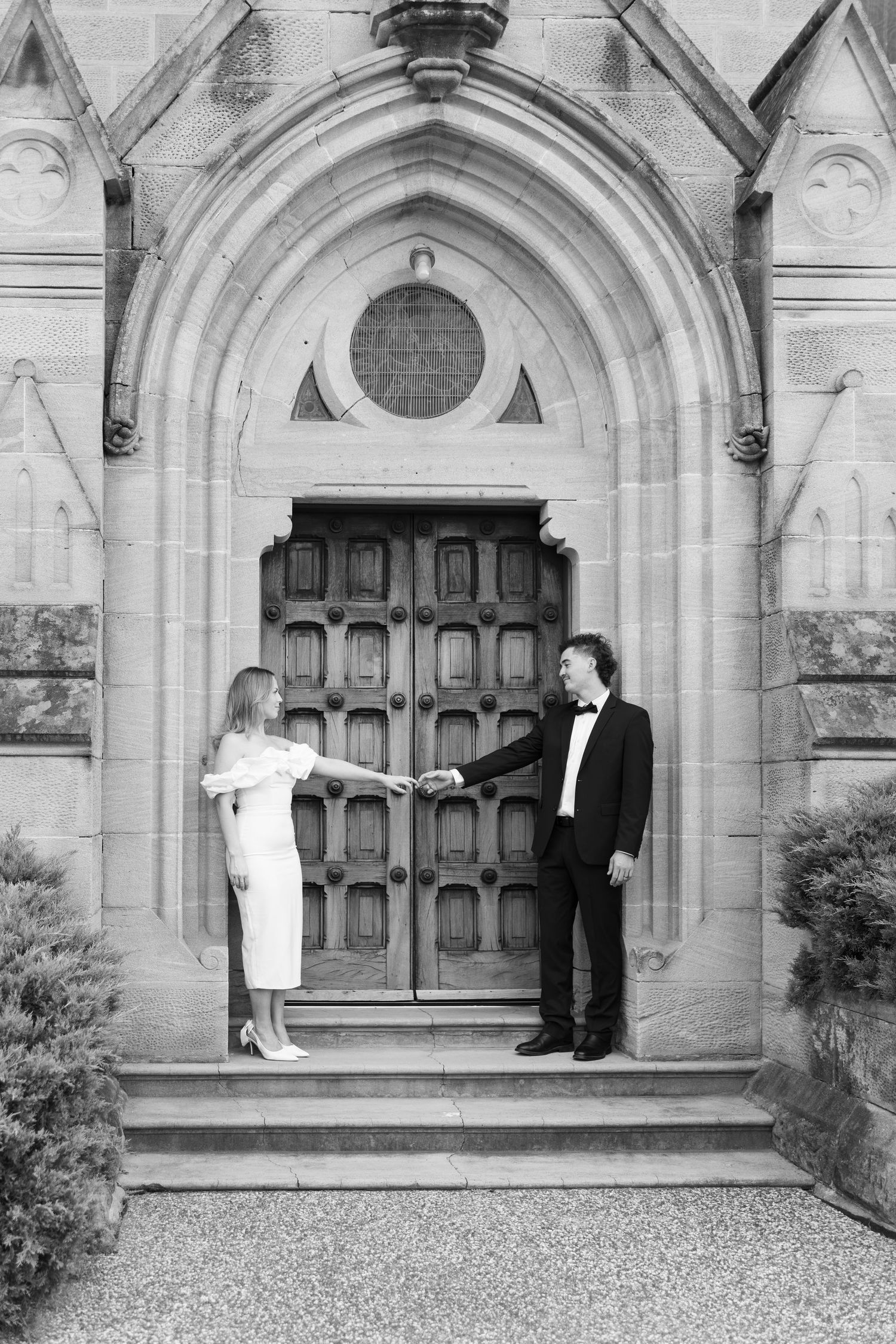 Black and white photo of elegant couple holding hands before gothic church doors in formal attire.