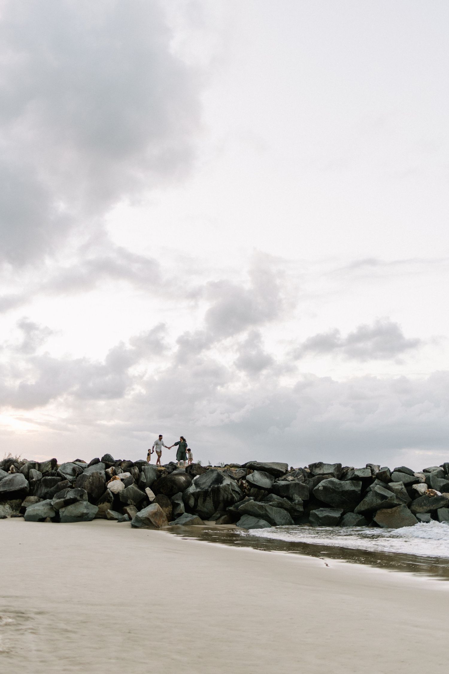 A family silhouetted on a rocky jetty by the ocean under a dramatic cloudy sky.