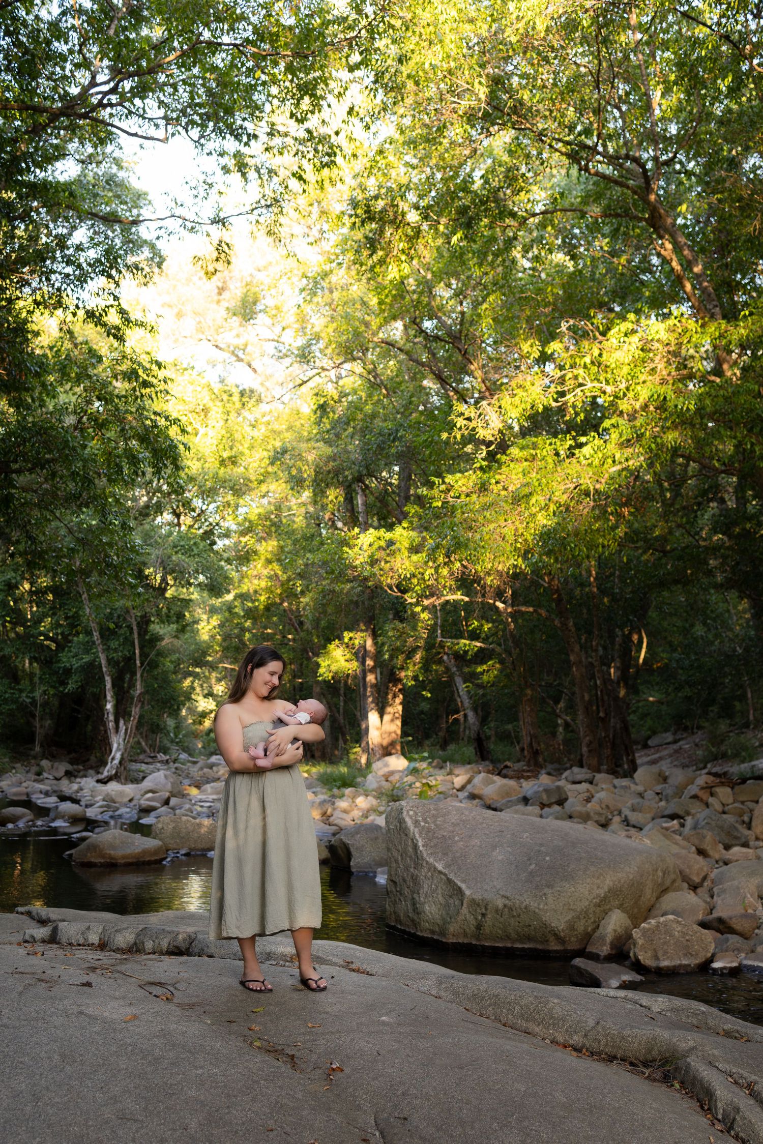 Mother holding newborn baby on rocky riverbank surrounded by lush green trees in natural outdoor setting.