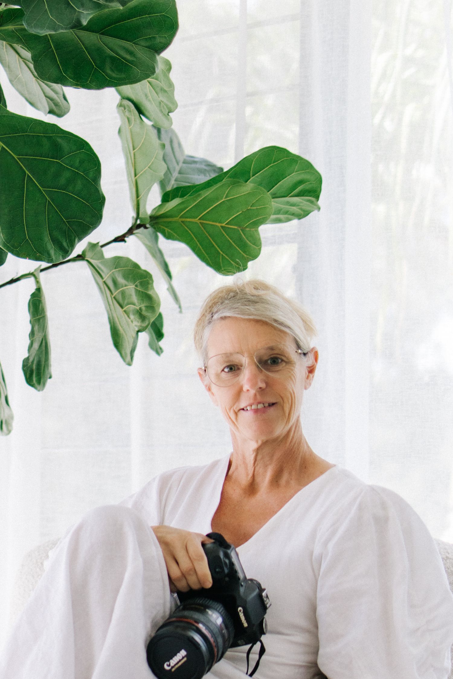 Smiling woman with short gray hair holding a Canon camera, seated near a fiddle leaf fig plant by a bright white window.