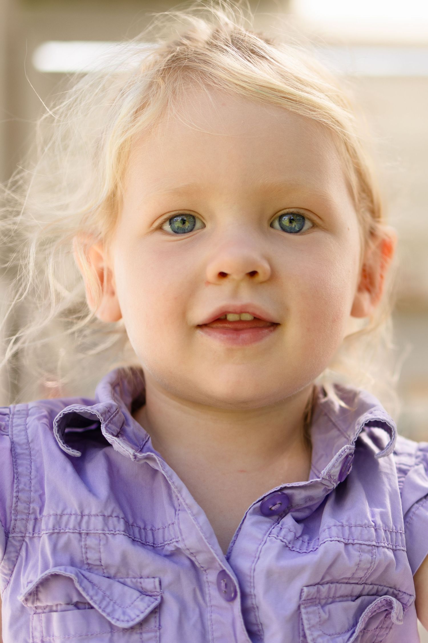 Adorable blonde toddler girl with blue eyes wearing a purple button-up shirt, smiling softly outdoors.