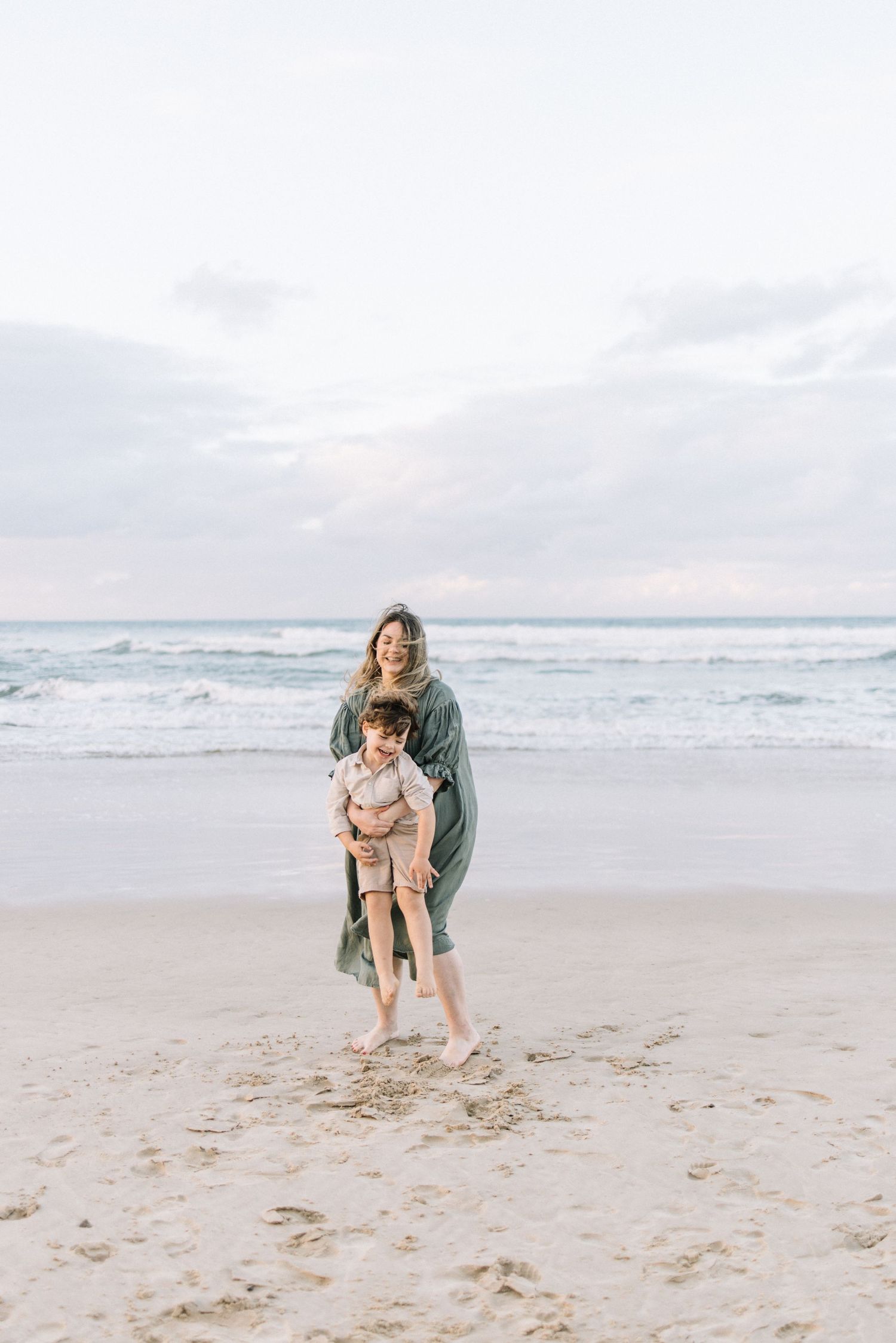 A smiling mother in a green dress playfully holds her child on a sandy beach.