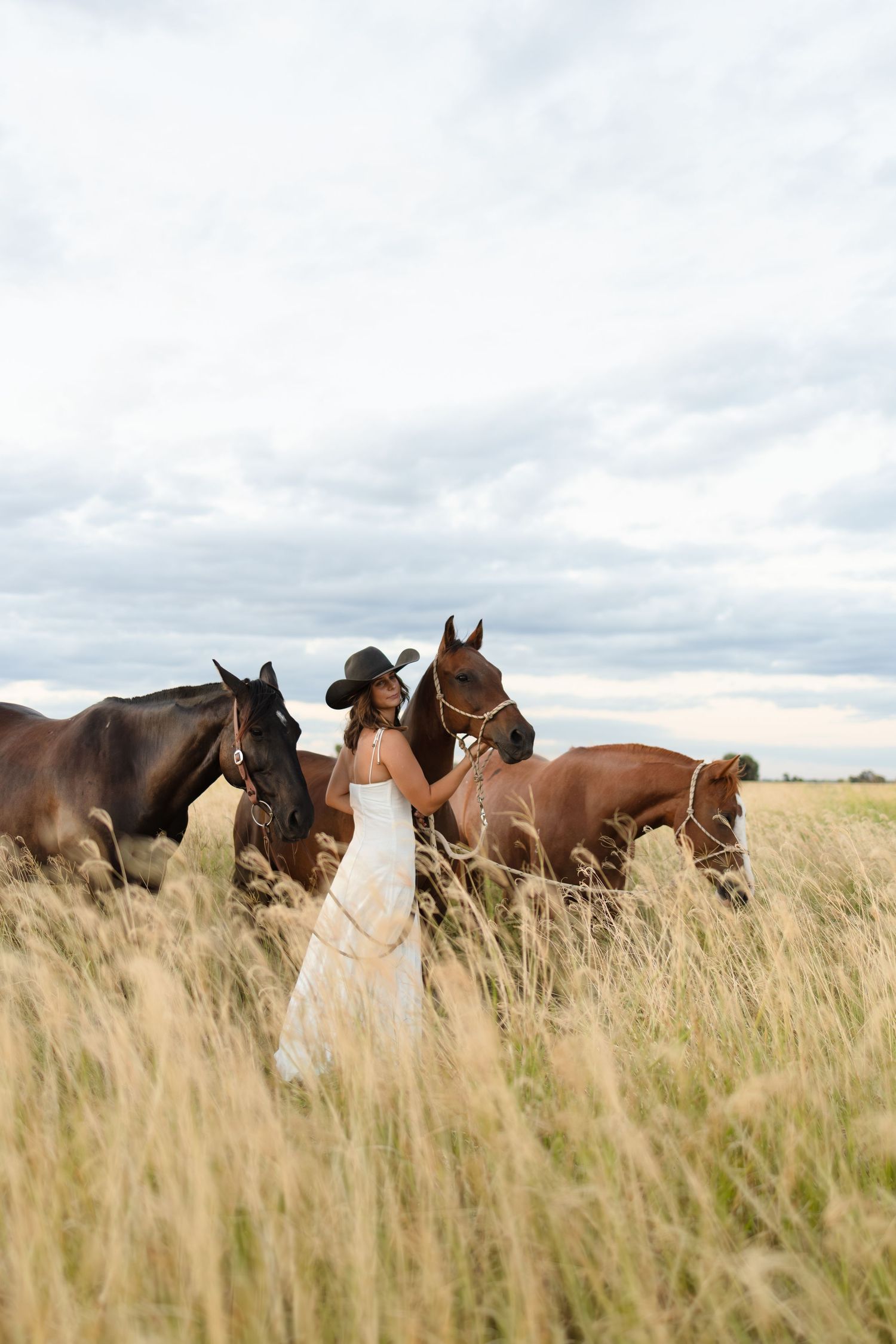 Woman in white dress and cowboy hat walks with two horses through tall golden grass in open field.