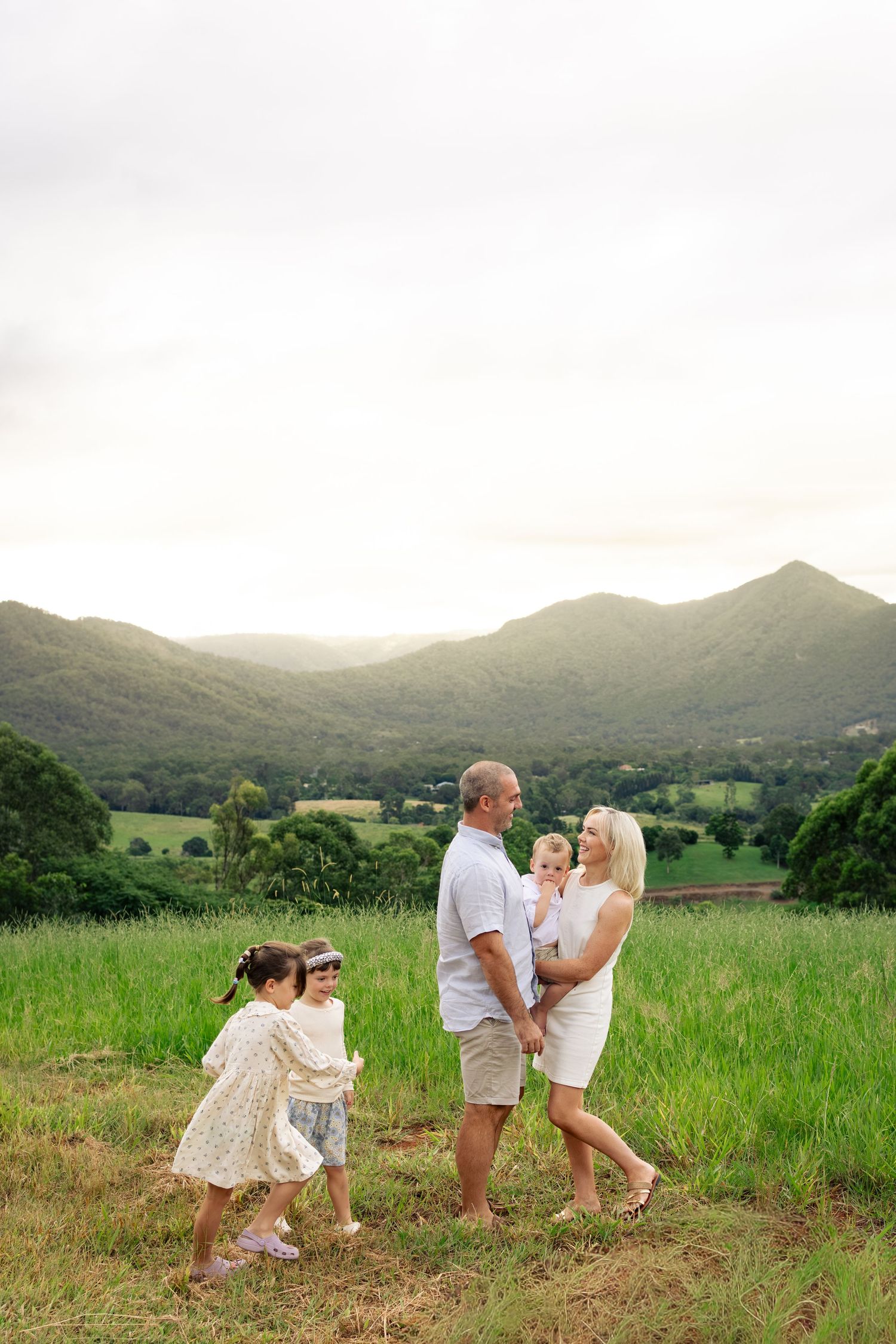 Family of four enjoying a sunny day in a lush green mountain meadow, parents holding children in casual white outfits.