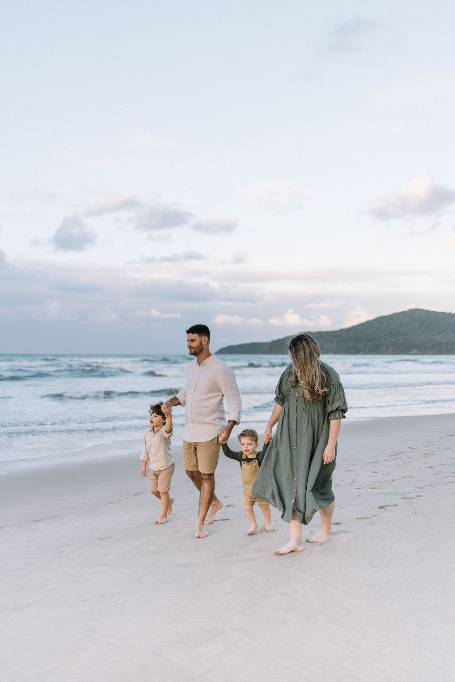 A family of four walks hand-in-hand along a sandy beach at dusk with ocean waves behind.