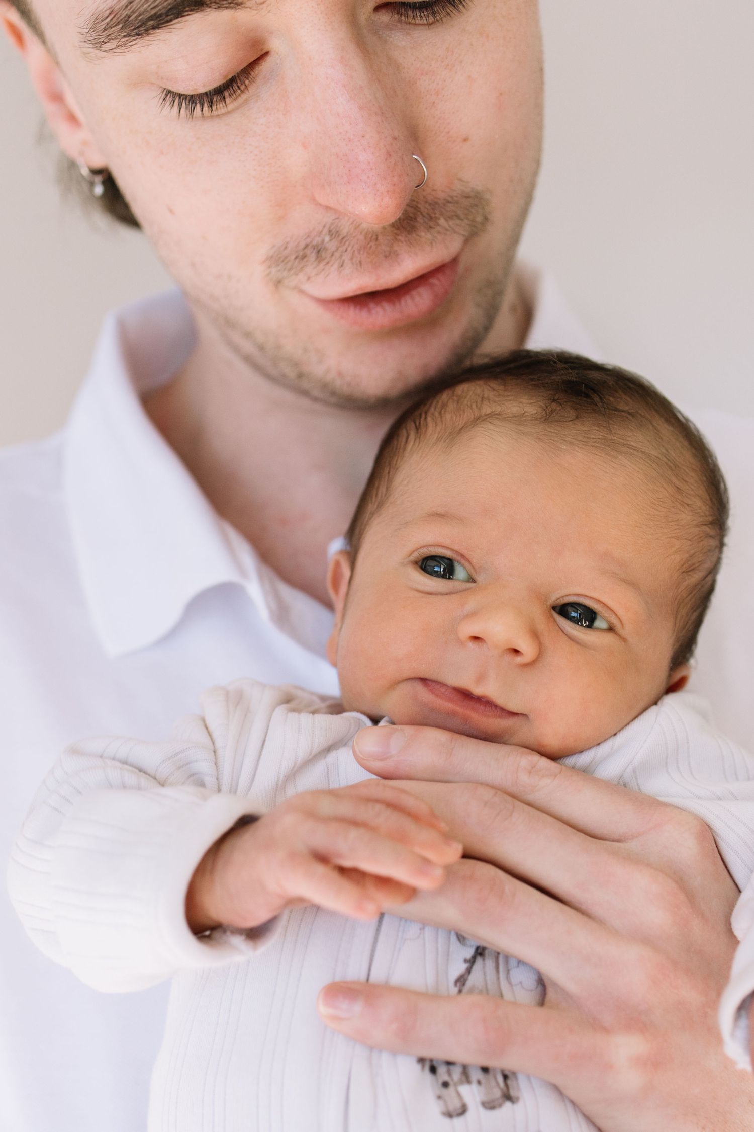 Young father with nose ring gently cradling his newborn baby in white clothing against light background.