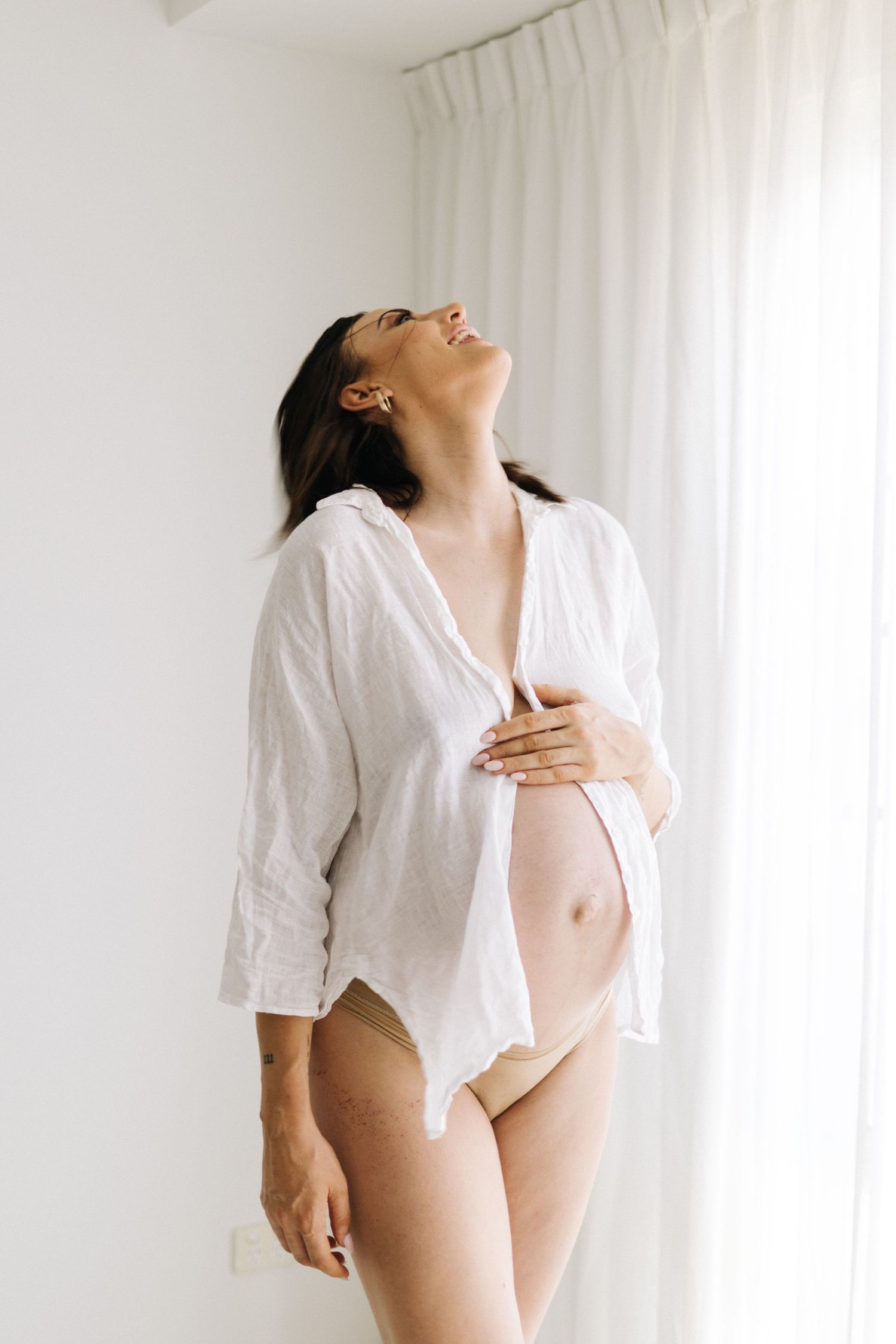 Pregnant woman in white linen shirt and beige underwear cradling baby bump, smiling with head tilted back.