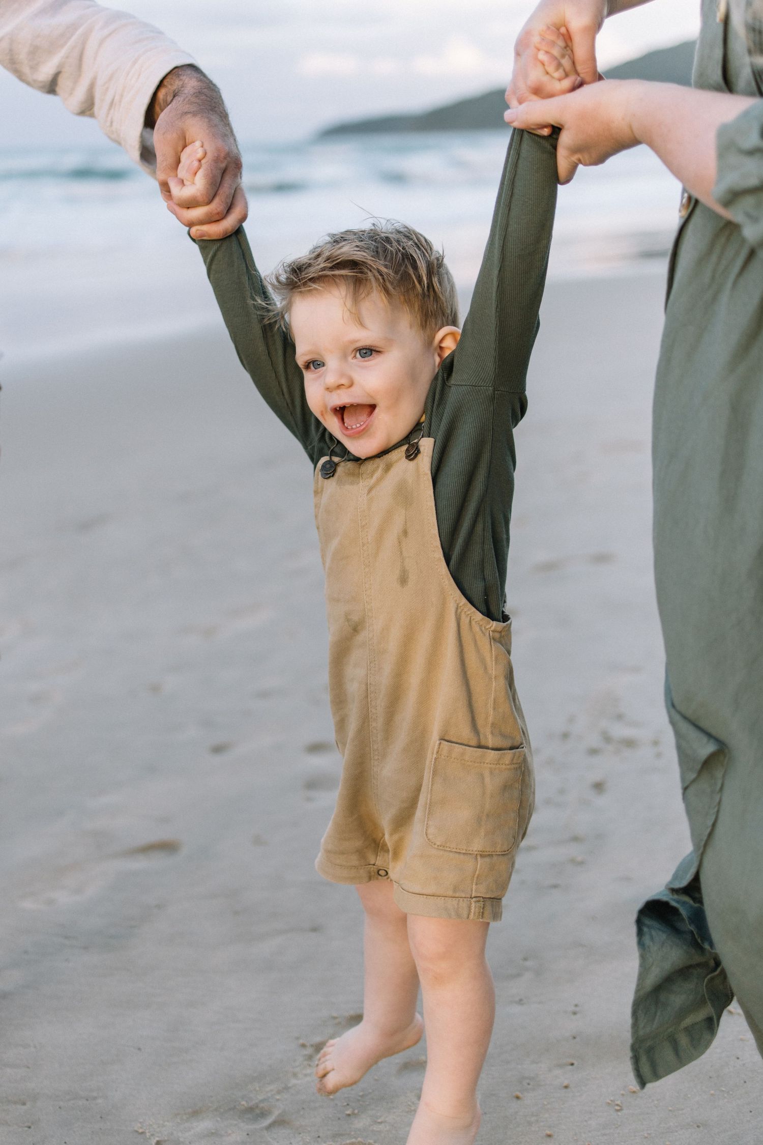 Joyful toddler in tan overalls holds adults hands on sandy beach, arms raised, laughing with ocean waves behind.