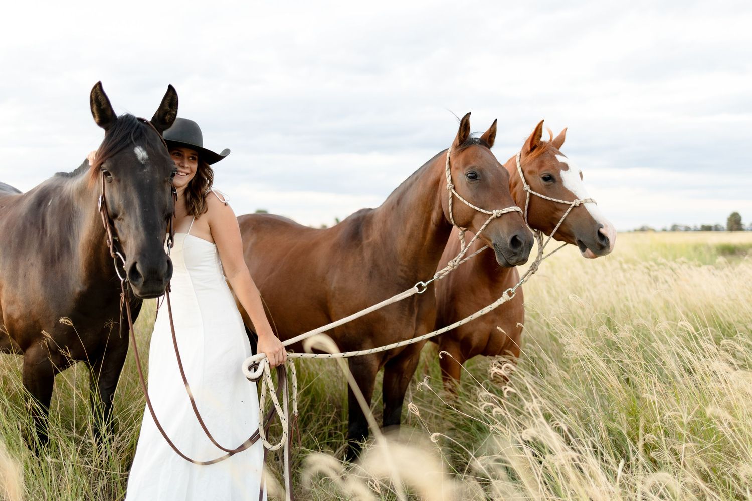 Woman in white dress and black hat stands with three horses in a golden field under overcast skies.