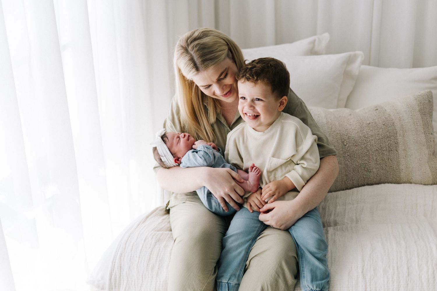 A smiling mother sits on a white bed holding her newborn baby while her young toddler son laughs beside her.