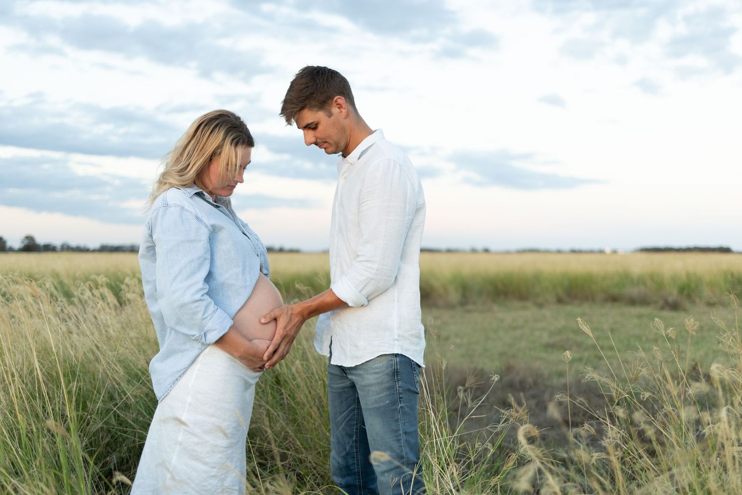 Couple looking down at pregnant belly while standing in golden grass field.