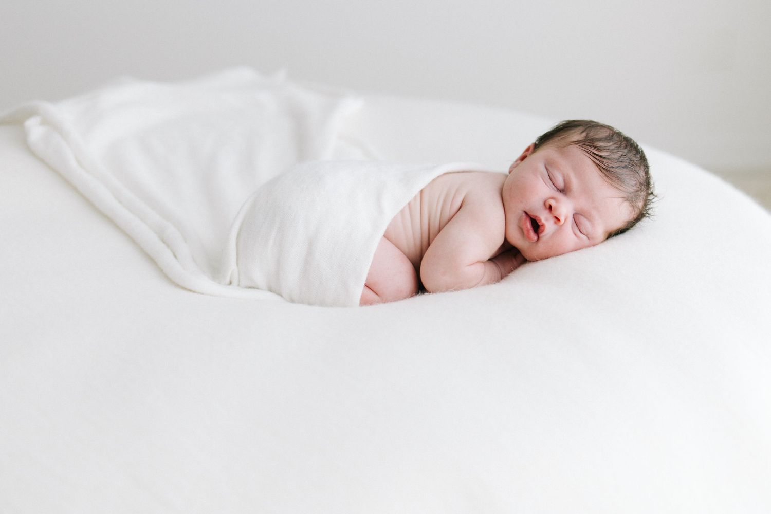 Newborn baby swaddled in white blanket sleeping on white rounded surface, mouth slightly open.