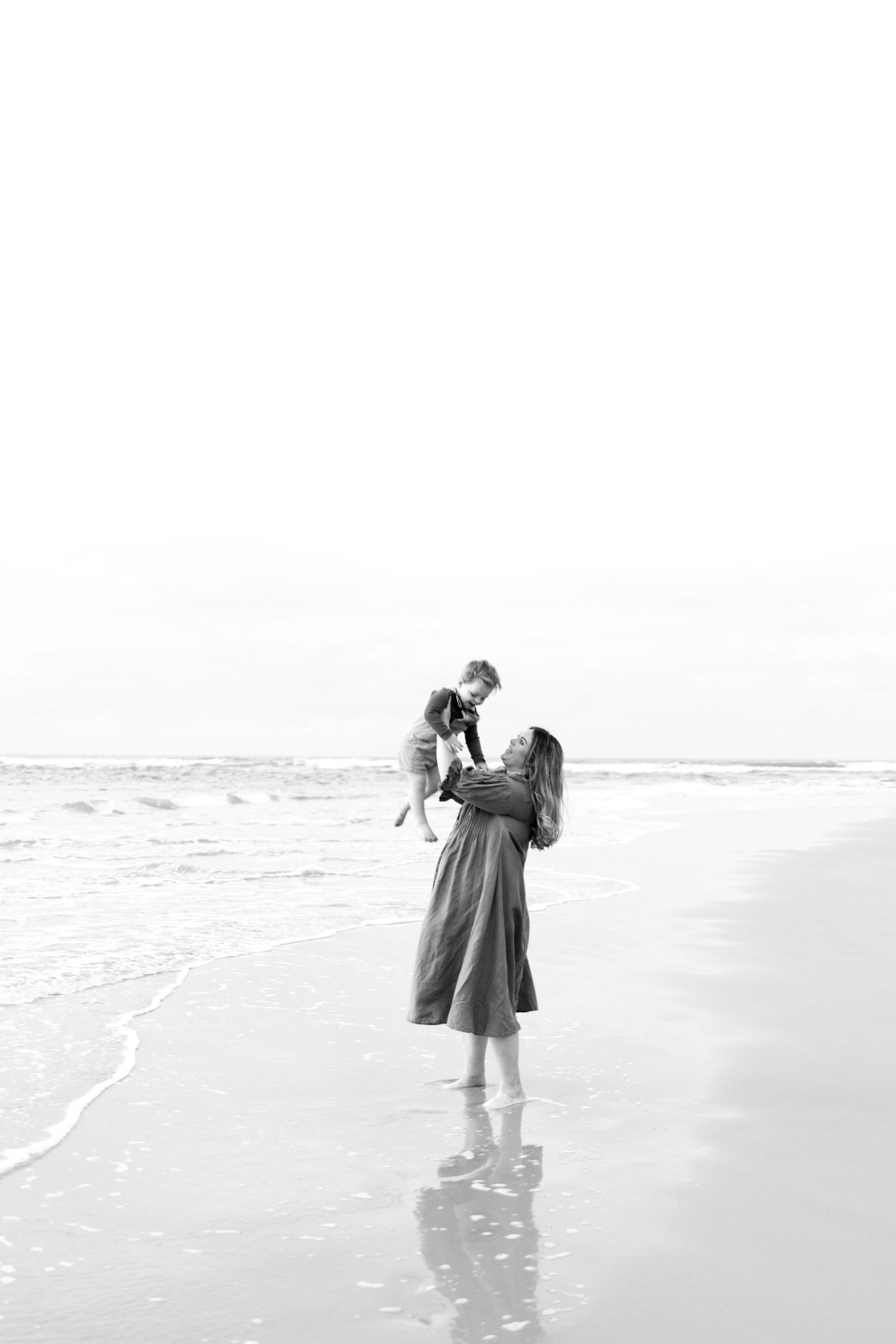 Black and white photo of a mother lifting her young child on a serene beach shoreline.