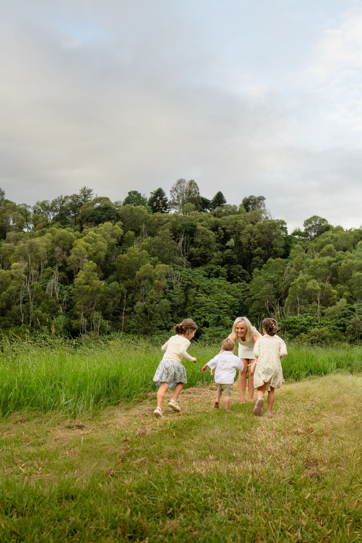 A mother and three children playing together in a lush green meadow with a forest backdrop on a cloudy day.
