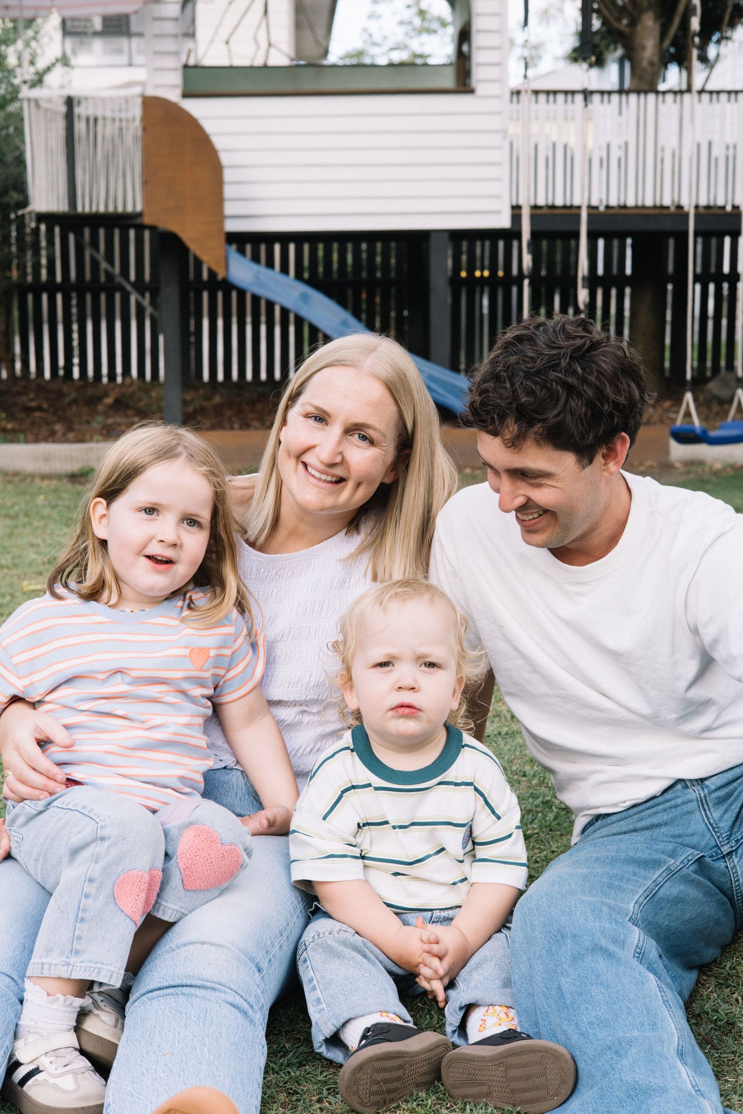 Happy family of four sitting on grass in backyard with playground, parents smiling at children on sunny day.
