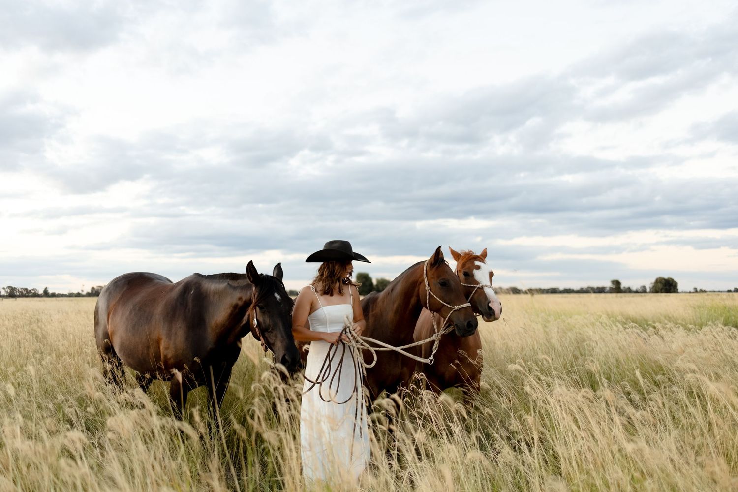 Woman in white dress and black cowboy hat leads two horses through a golden grass field under cloudy skies.