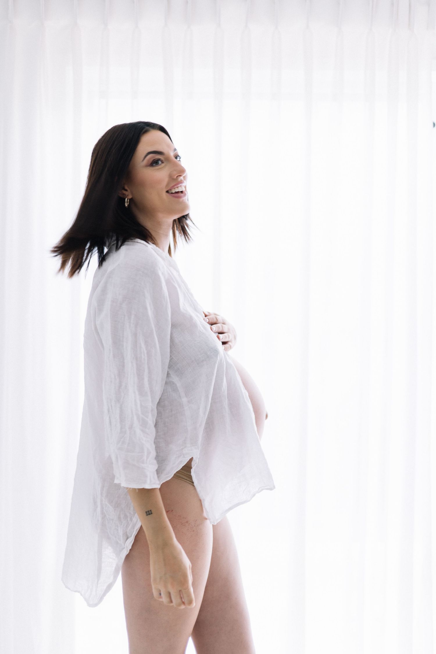 Smiling pregnant brunette in open white linen shirt standing before white curtains showing baby bump.