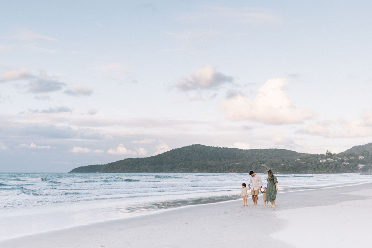 Family walking along a serene white sand beach at dusk with gentle waves and green hills in the background.